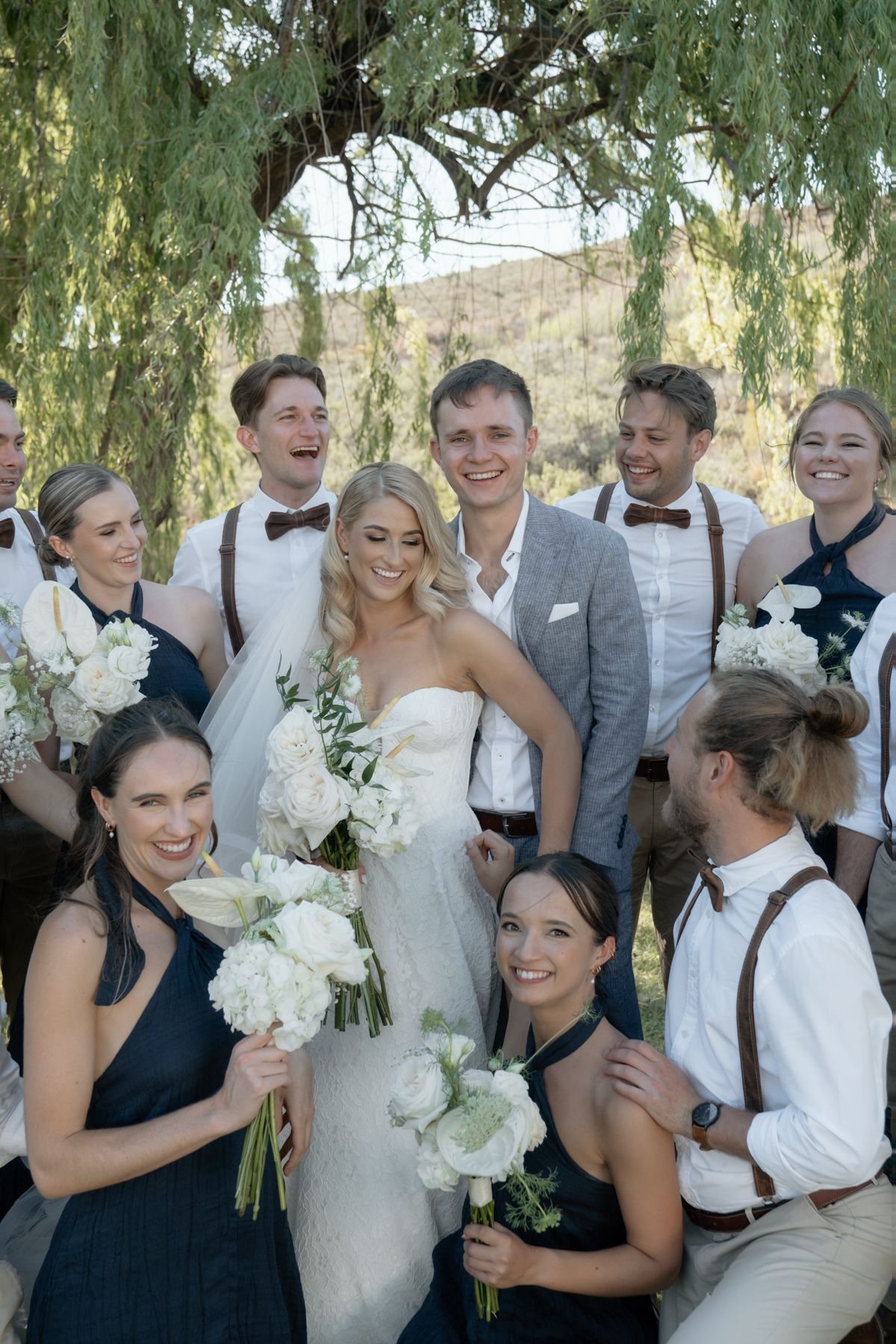 Wedding party poses under a weeping willow, bridesmaids in navy dresses with white bouquets and groomsmen in suspenders with bow ties.