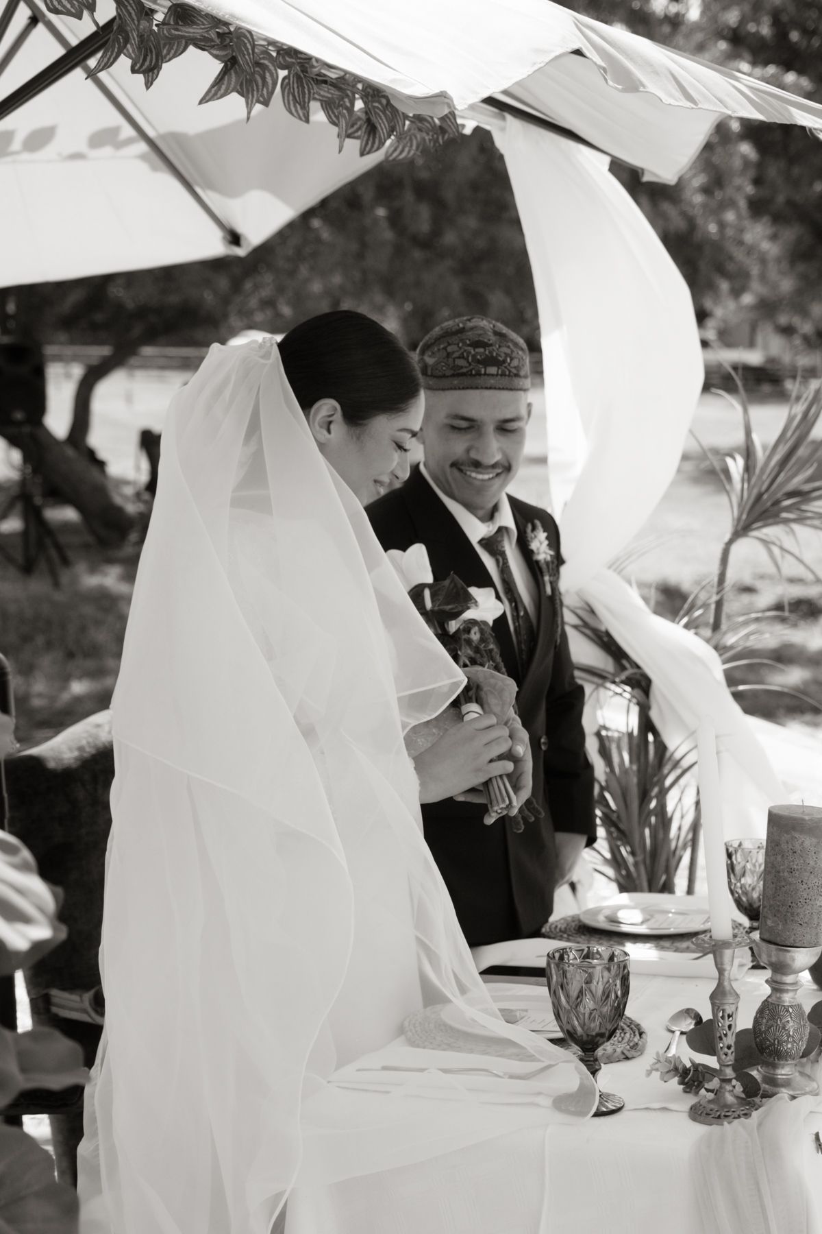 Black and white photo of smiling bride and groom under chuppah during Jewish outdoor wedding ceremony.