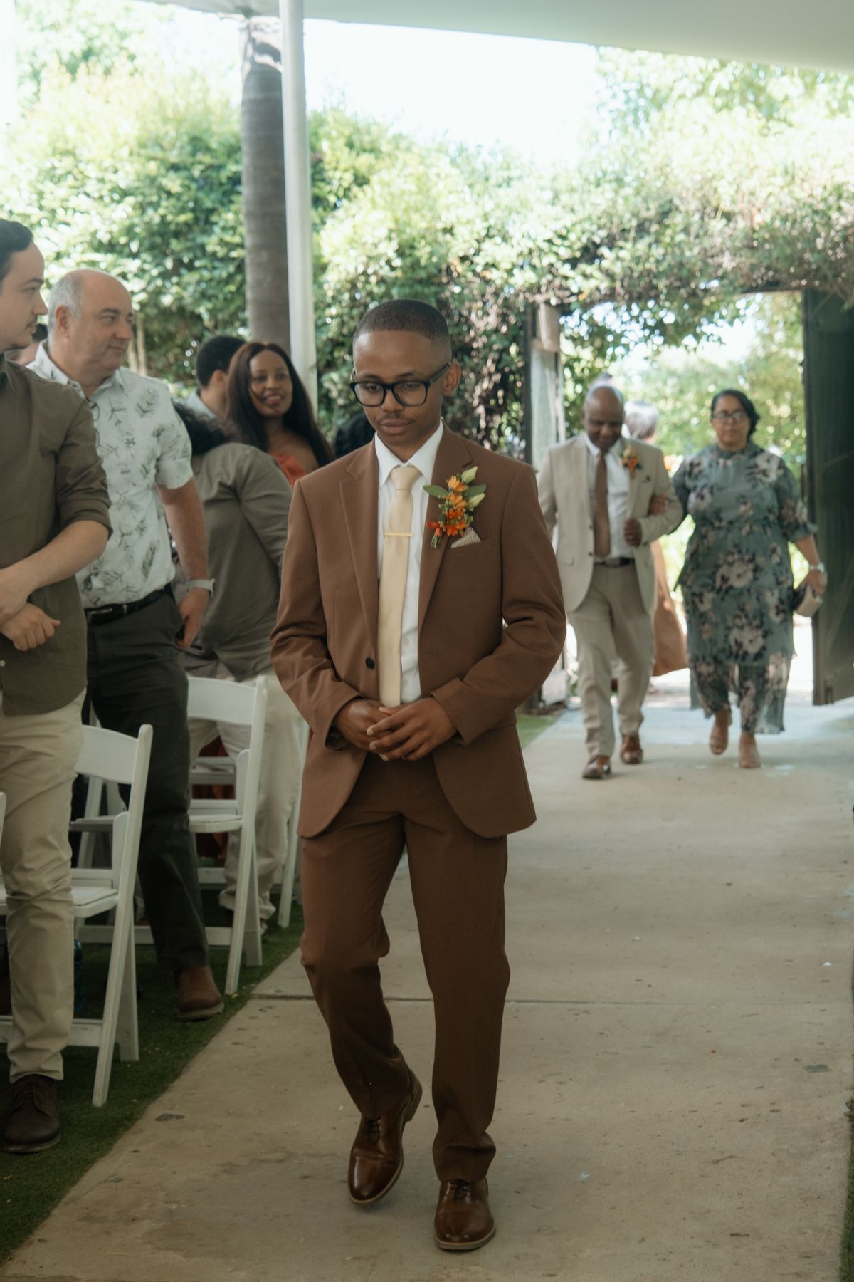 Groom in brown suit with floral boutonniere walks down aisle at outdoor garden wedding ceremony.