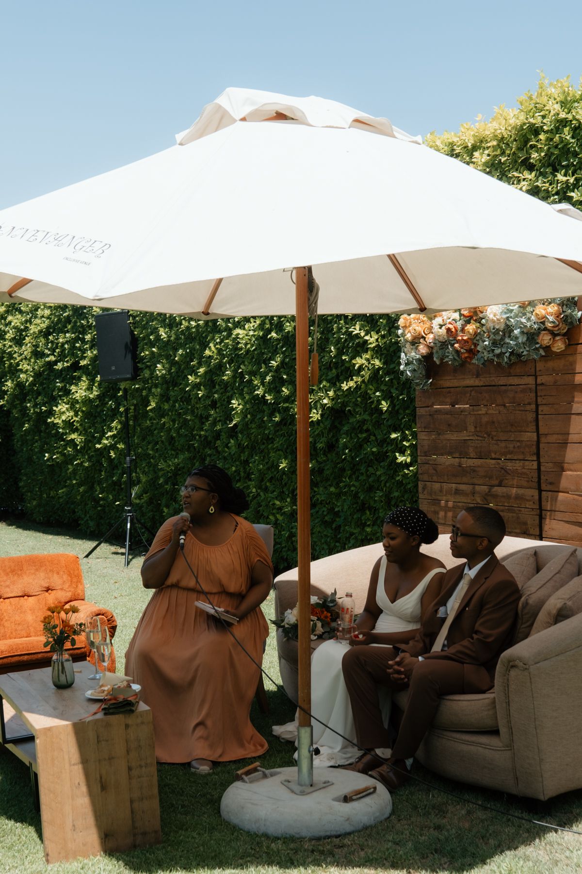 A woman in an orange dress speaks at an outdoor wedding reception while the couple sits on a sofa under a white umbrella.