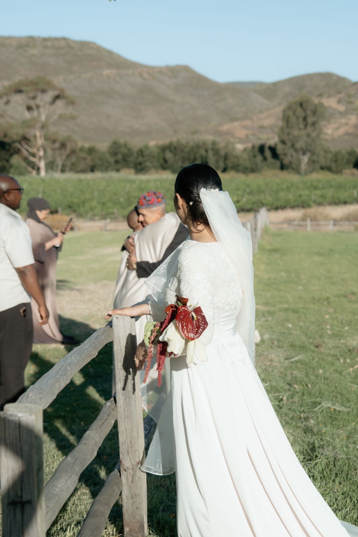Bride in white wedding gown with veil holding bouquet near wooden fence at outdoor rural ceremony.