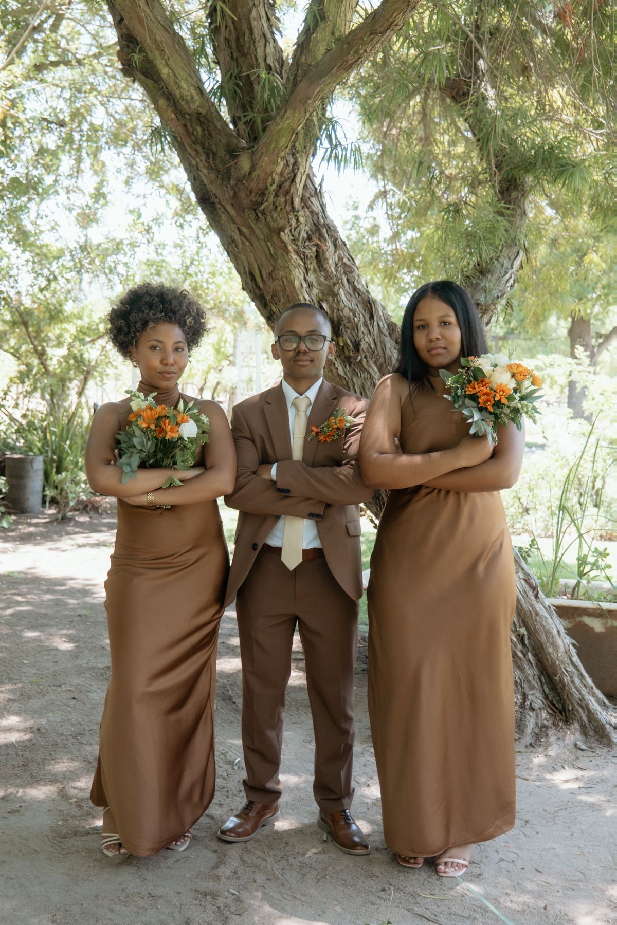 Groom in brown suit poses with two bridesmaids in brown gowns holding orange bouquets with arms crossed outdoors.