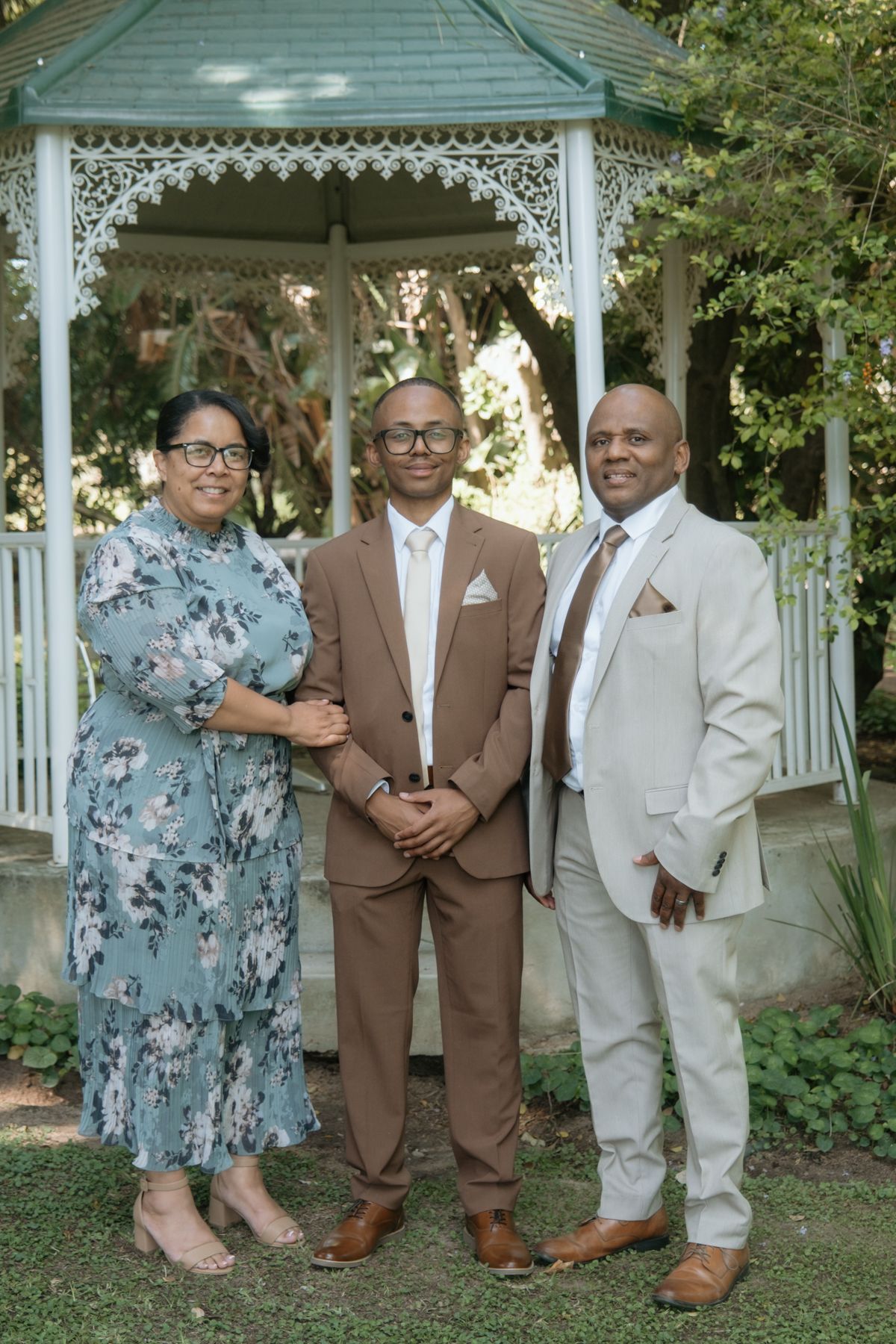 A family of three poses in front of a white gazebo, with the son in a brown suit flanked by his parents.