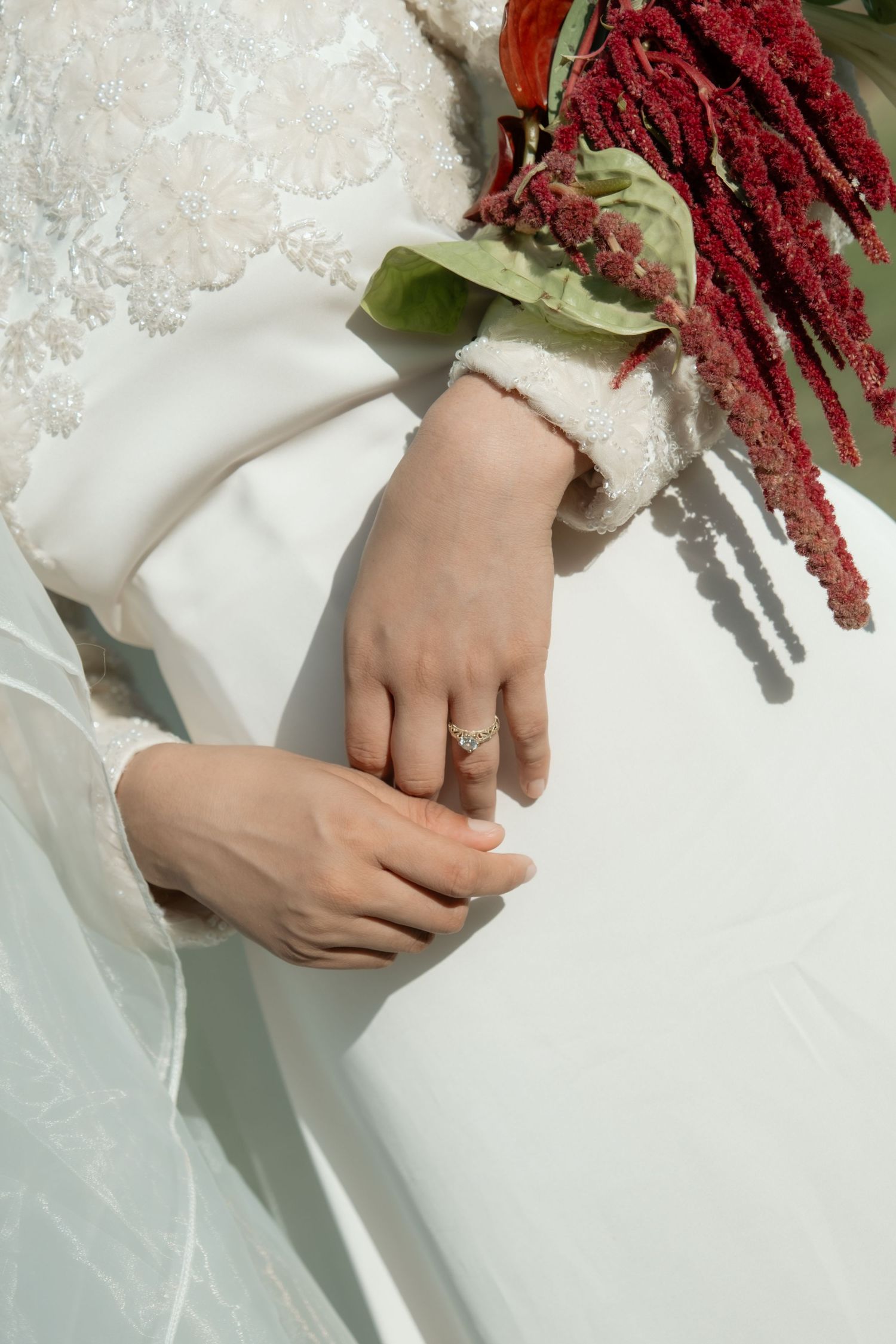 Bride in white lace wedding dress holding red amaranth bouquet with diamond ring on finger.