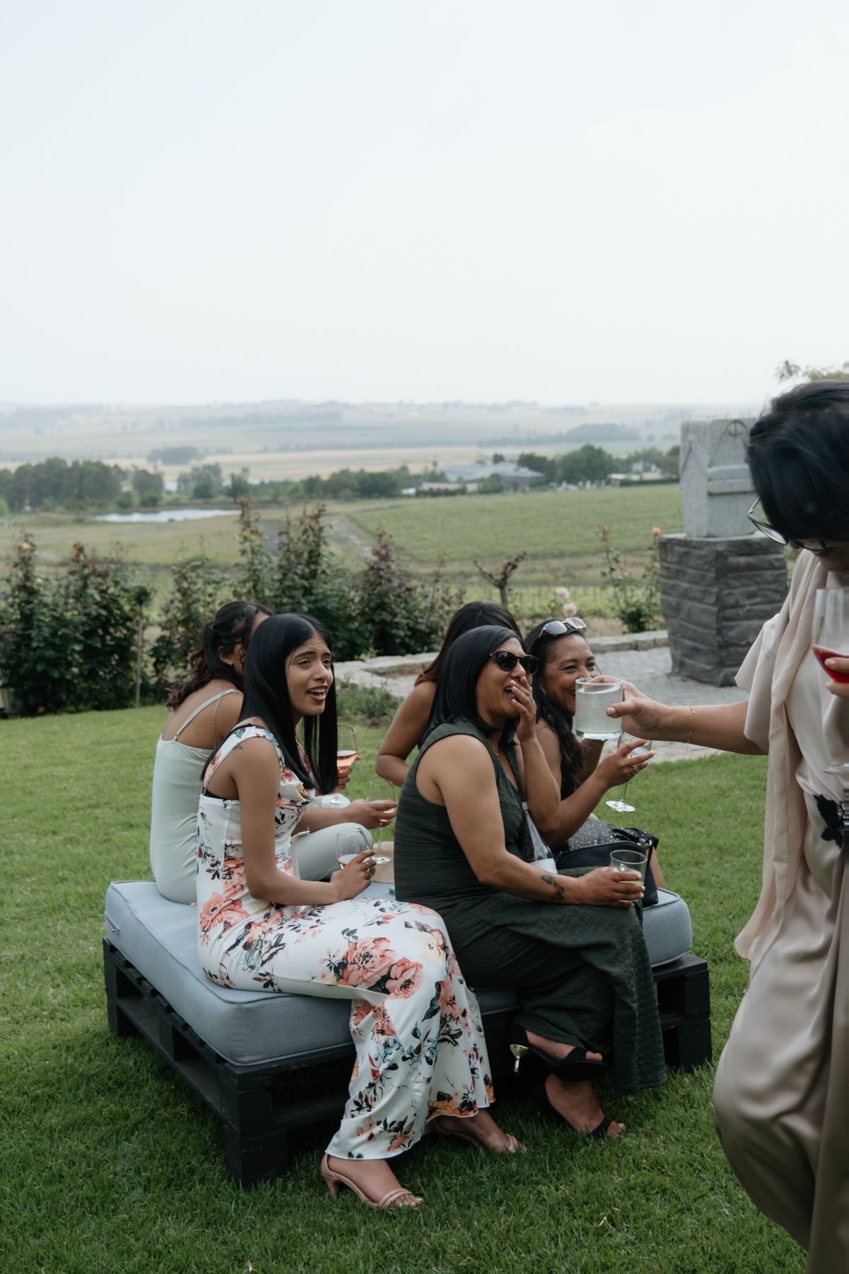 Group of women laughing and socializing outdoors on a pallet bench with scenic vineyard views.
