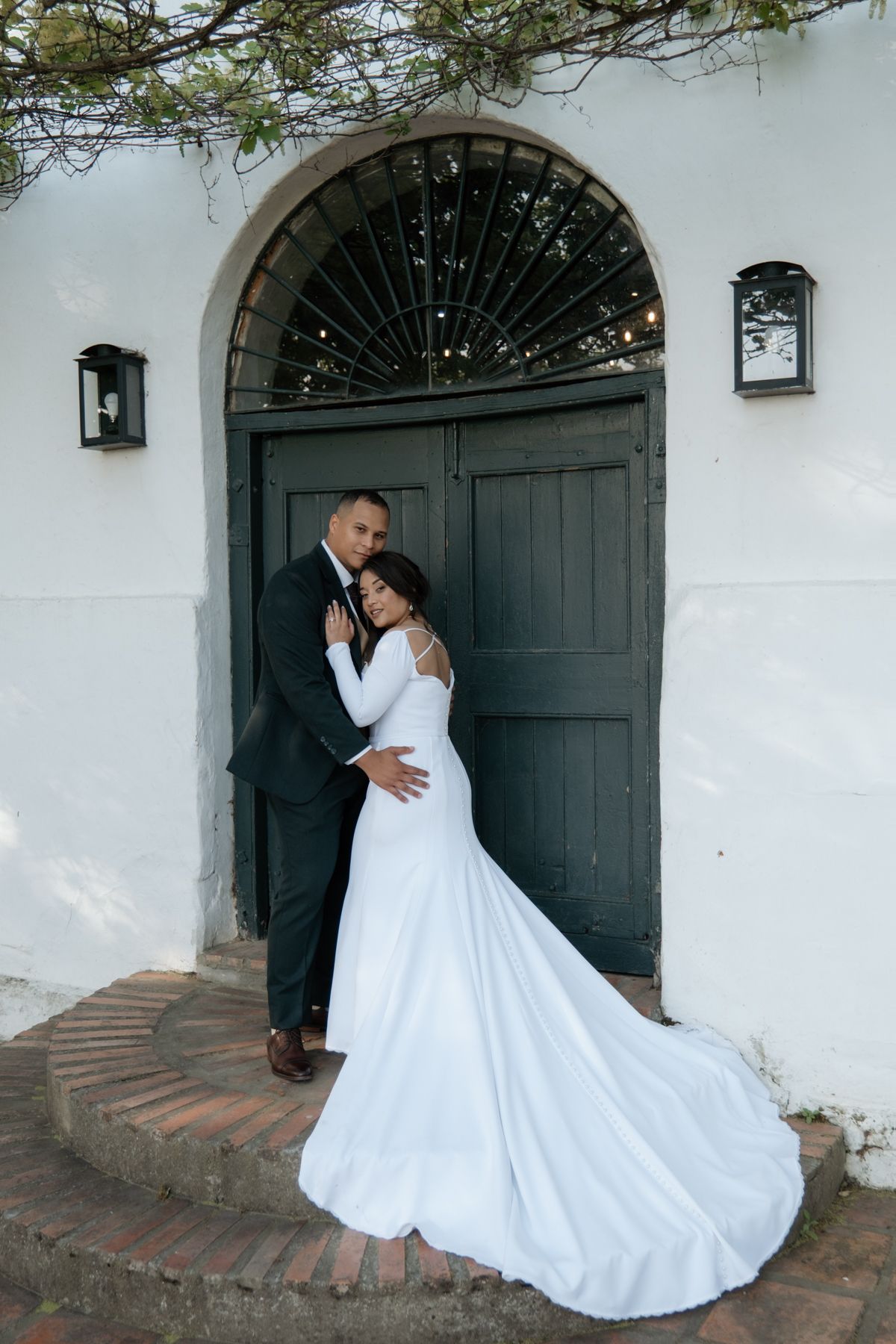 Bride and groom posing elegantly before arched dark wooden door of white historic building with trailing gown.