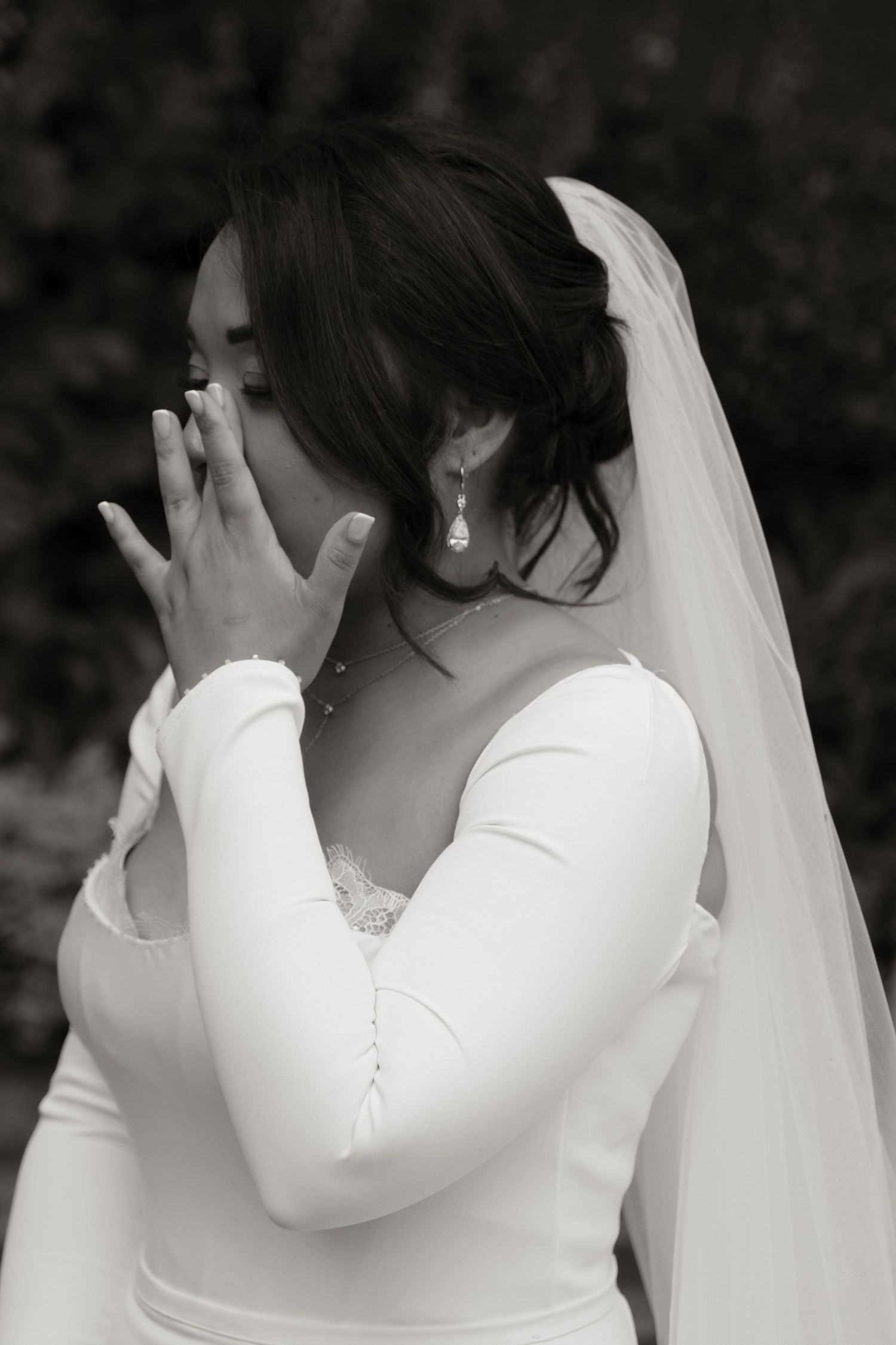 Emotional bride in white gown and veil covering her face with hands in black and white photo.