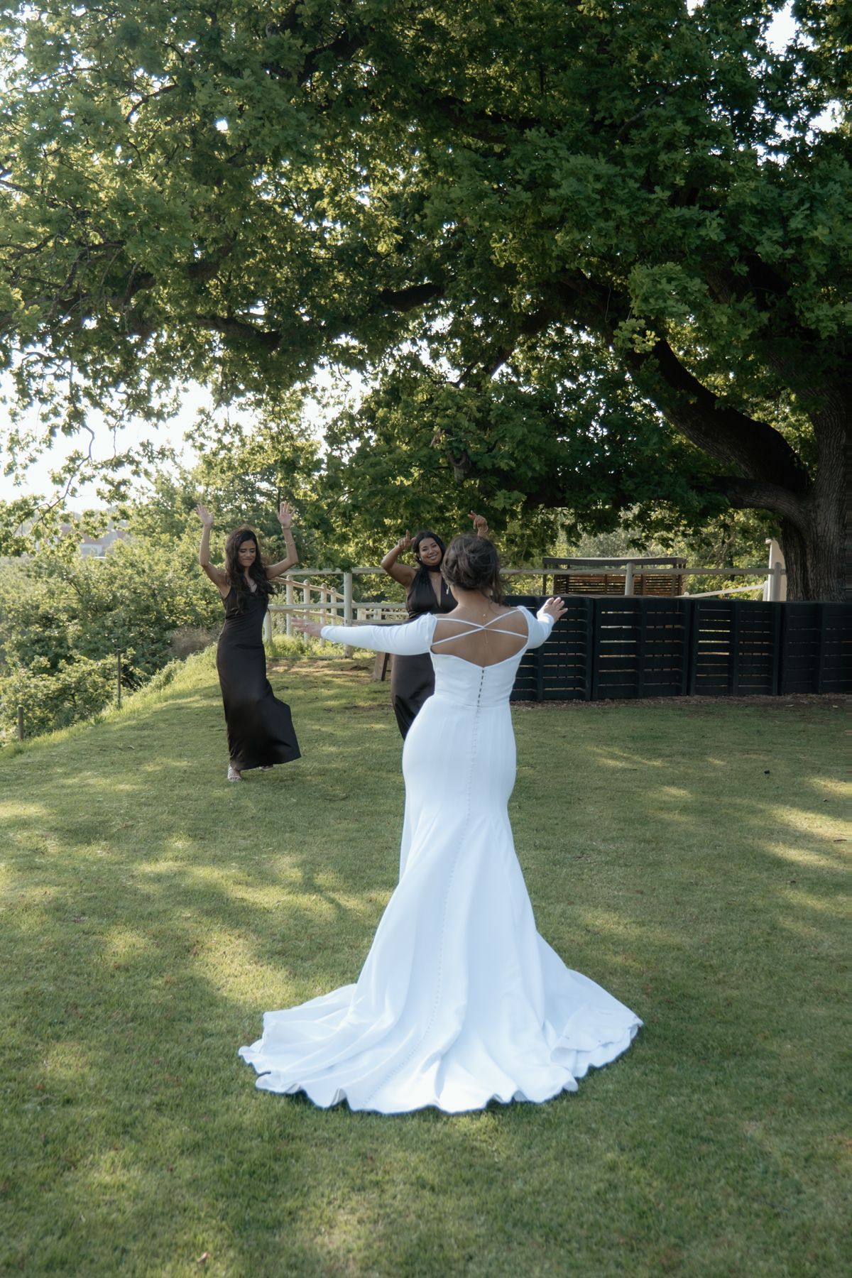 Bride in backless white gown with train opens arms to greet guests on a lush green lawn.