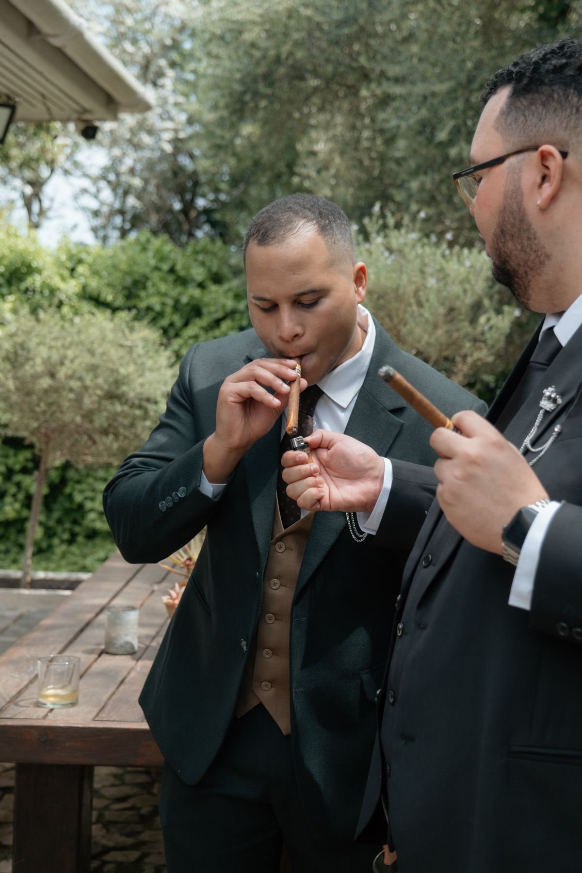 Groom in dark suit and vest lights a cigar outdoors with groomsman at wedding celebration.