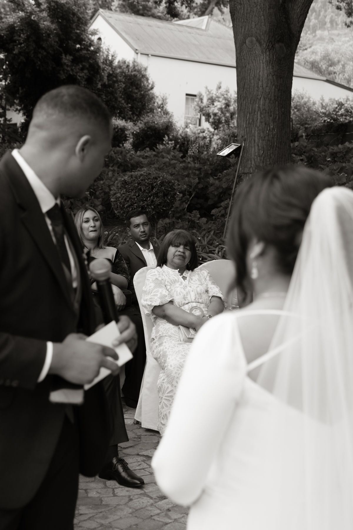 Black and white photo of groom reading vows to bride during outdoor wedding ceremony with seated guests.