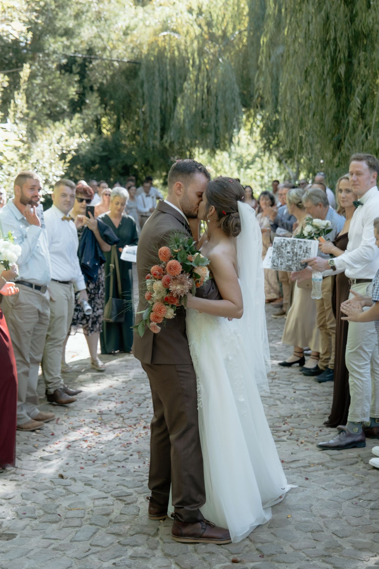 Bride and groom share first kiss as newlyweds surrounded by cheering guests on tree-lined path.