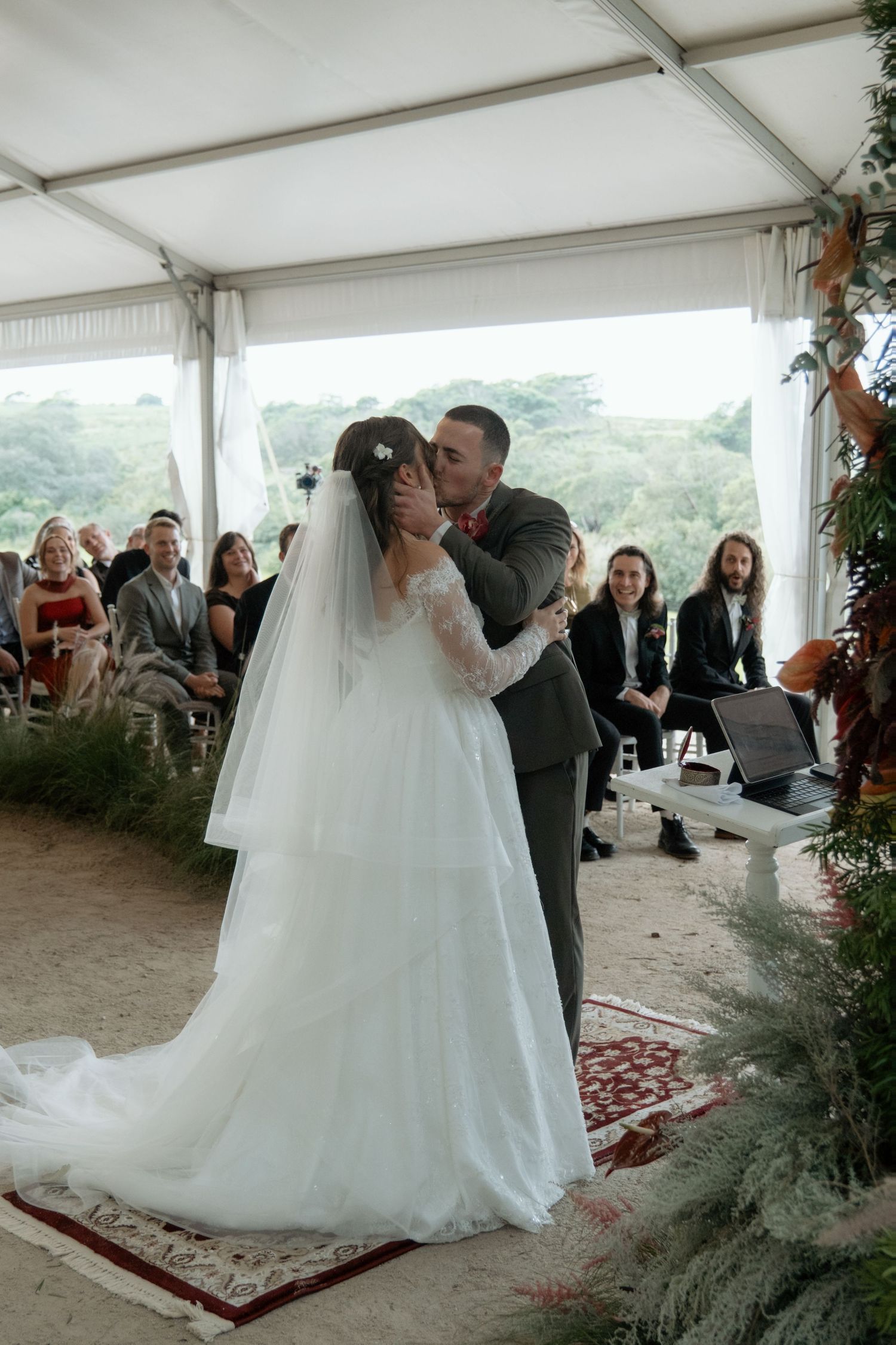 Bride and groom share first kiss at outdoor wedding ceremony under white tent with guests watching.