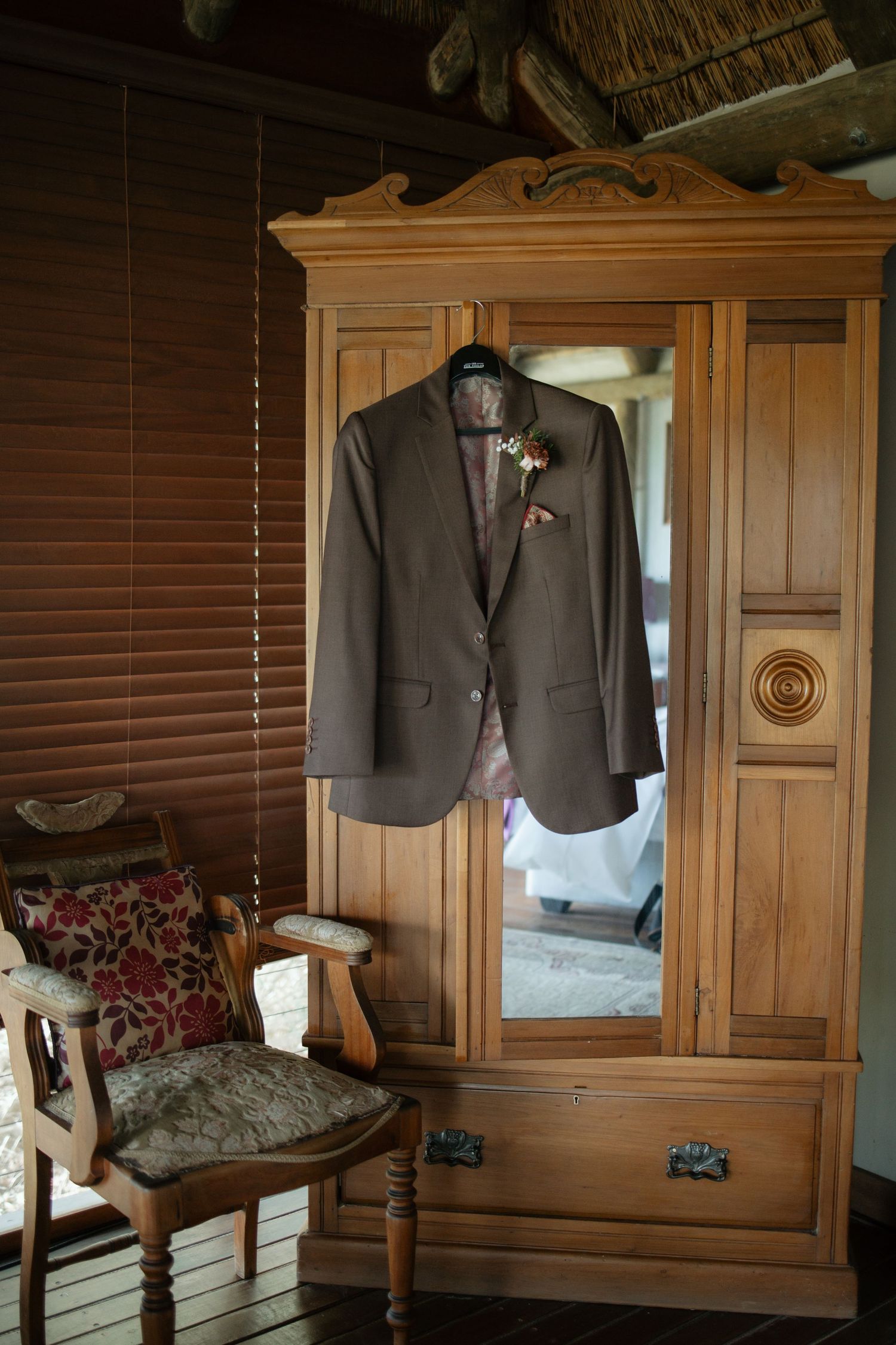A groom's brown suit jacket with boutonniere hangs on a wooden armoire in a rustic wedding venue.