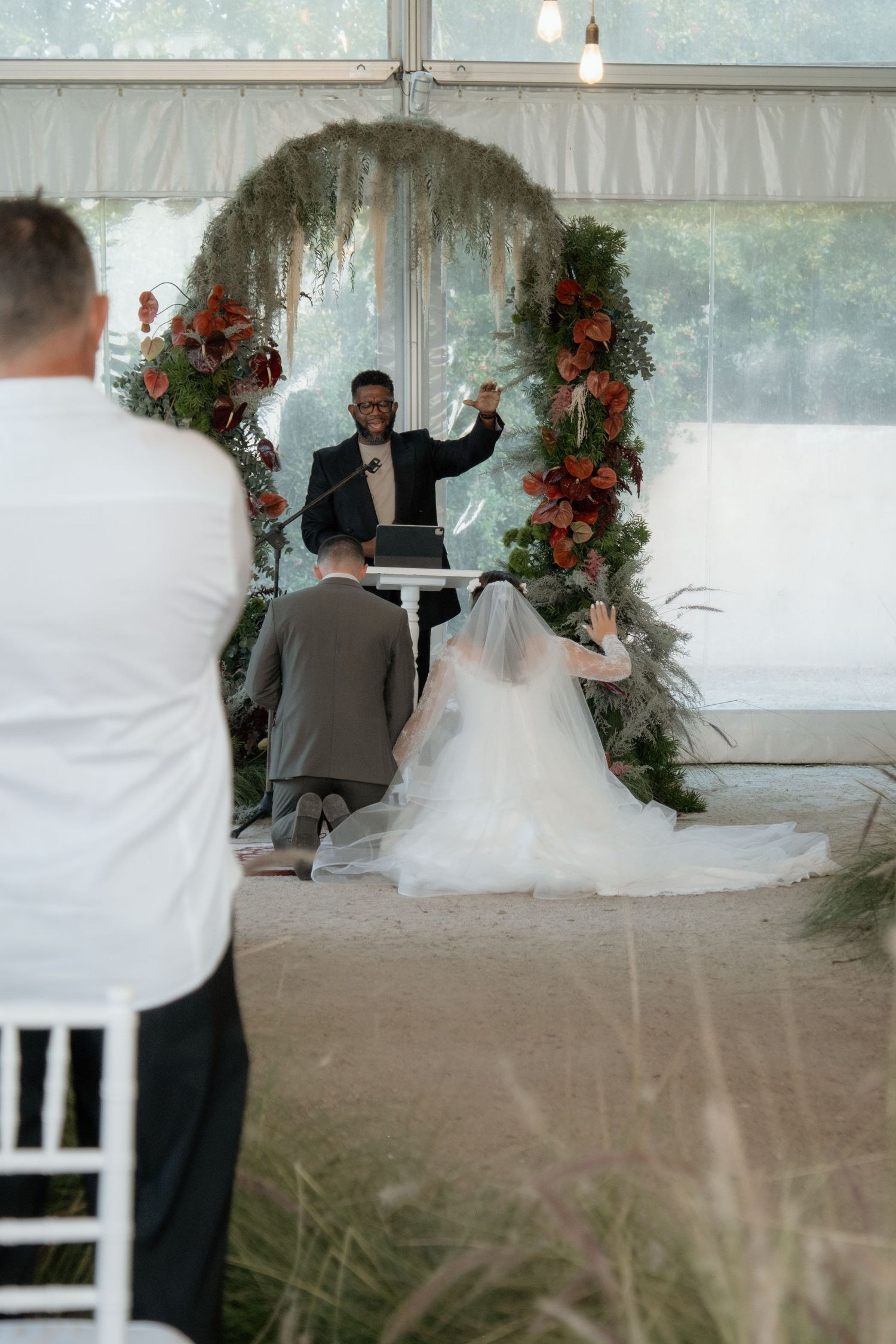 Officiant raises hand during indoor wedding ceremony with couple kneeling before floral arch adorned with greenery and red flowers.
