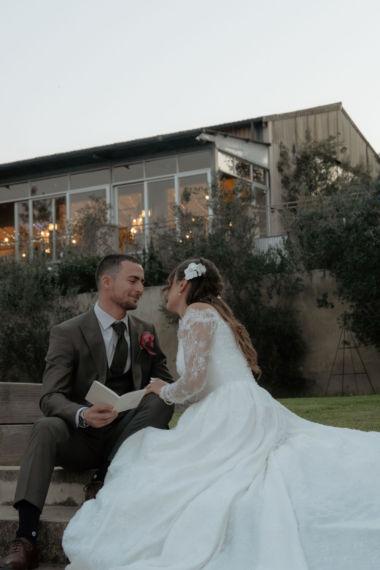 Groom reads from book while bride smiles at him on stone steps outside lit wedding venue at dusk.