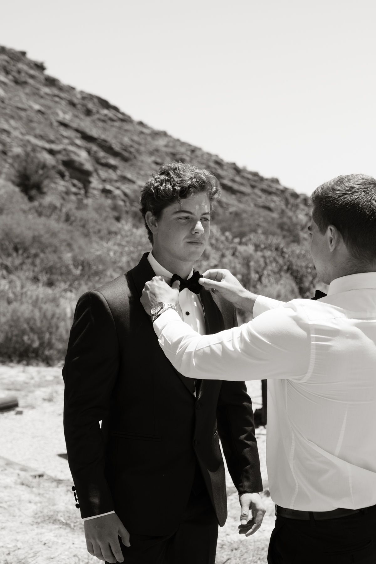 Black and white photo of a man in a tuxedo having his bow tie adjusted outdoors.