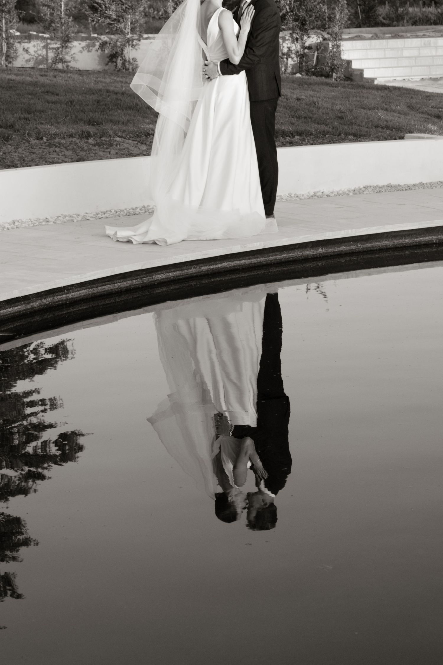 Black and white reflection of bride and groom kissing beside a still pool of water.