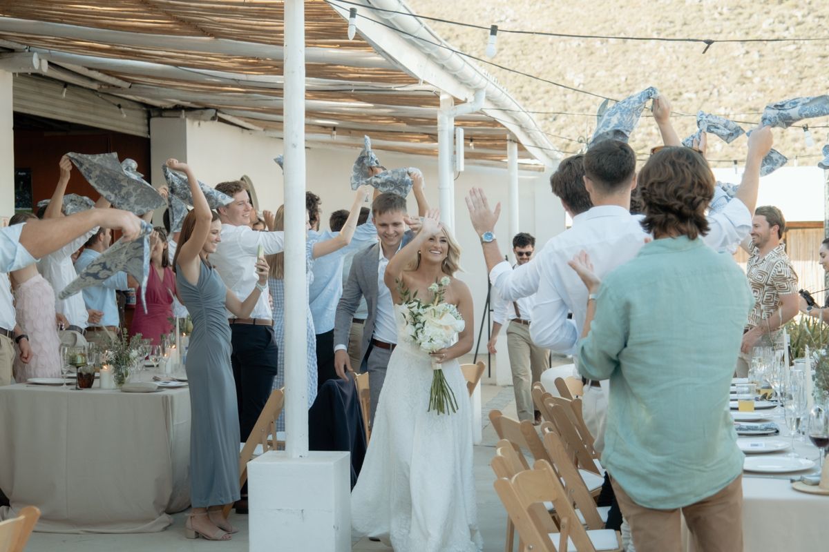 Bride and groom walk through cheering guests waving fabric at outdoor wedding reception celebration.