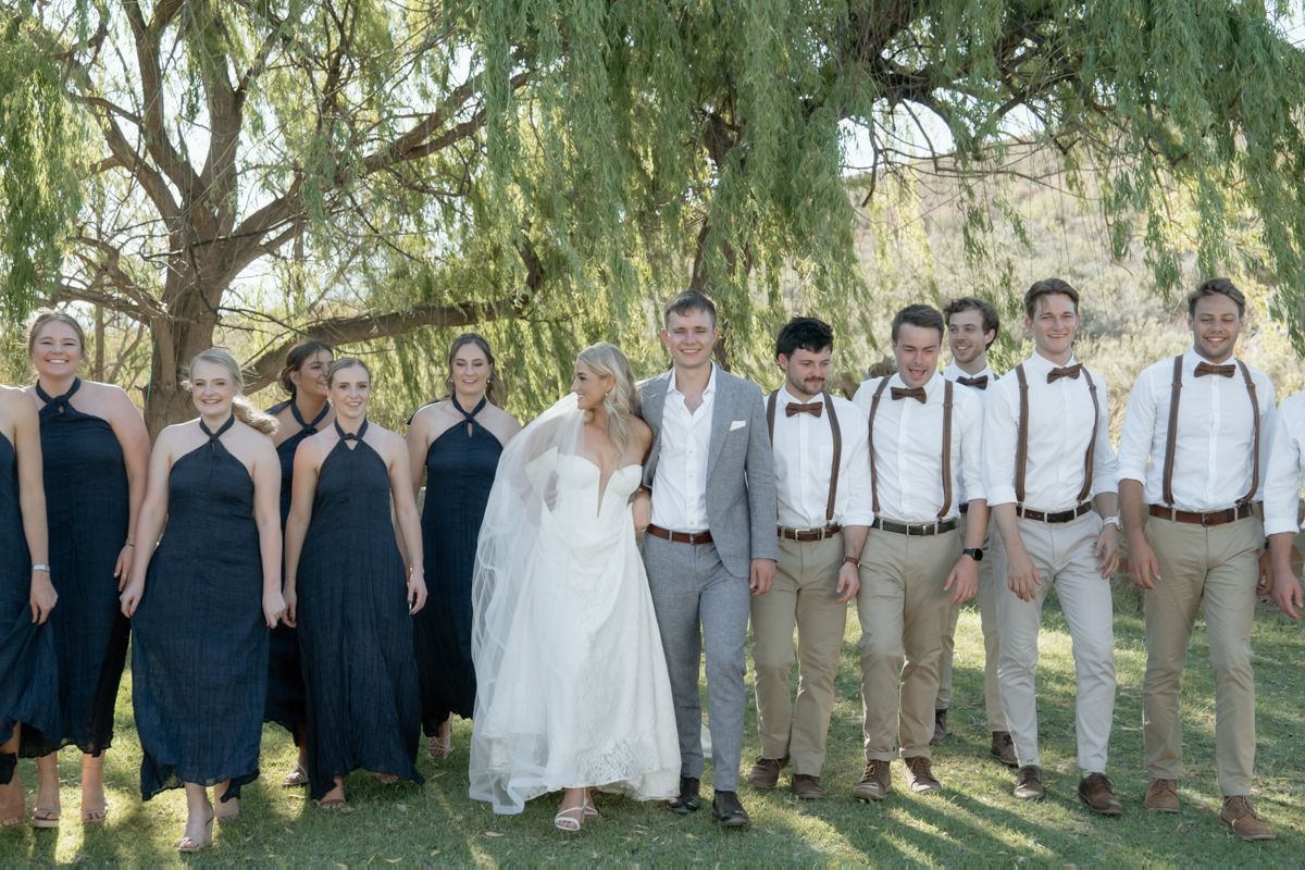 Wedding party posing together under a willow tree with bridesmaids in navy gowns and groomsmen wearing suspenders.