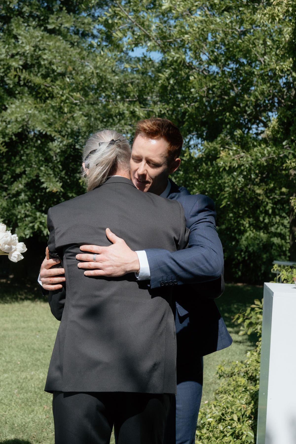 Groom in blue suit shares a warm embrace with an older gentleman during an outdoor wedding ceremony.
