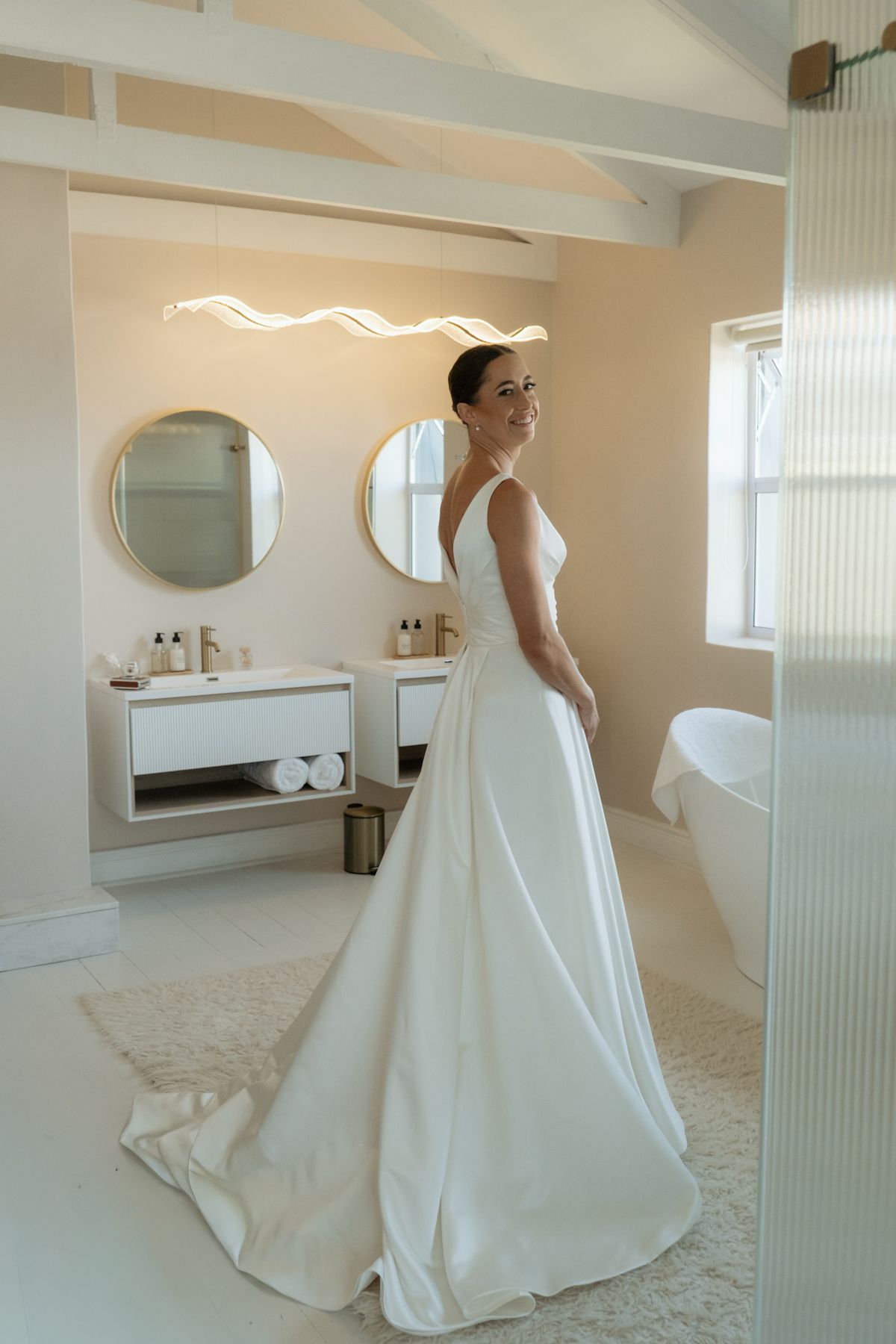 Radiant bride in simple white gown standing in modern luxury bathroom with gold-framed mirrors.
