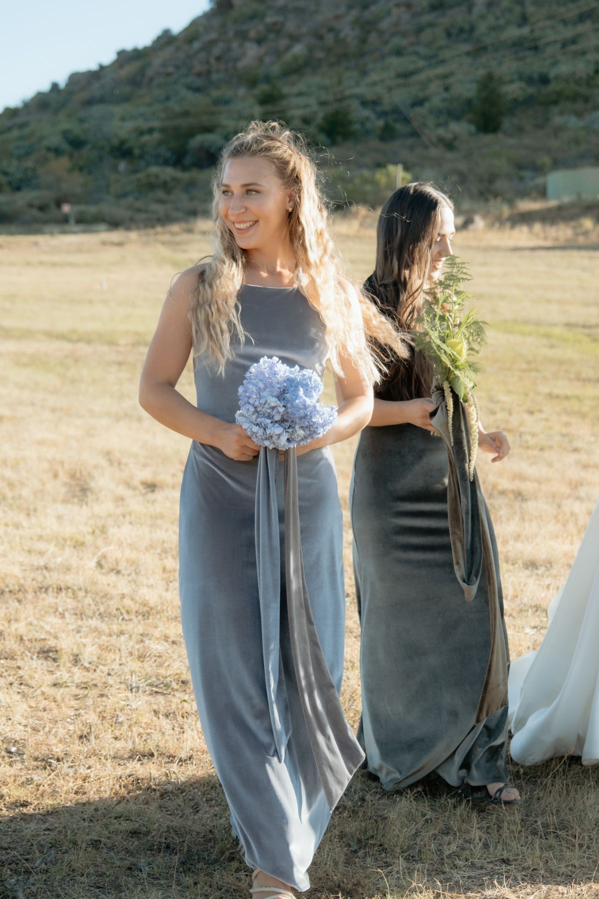 Smiling bridesmaid in grey velvet dress holding blue hydrangea bouquet at outdoor wedding ceremony.