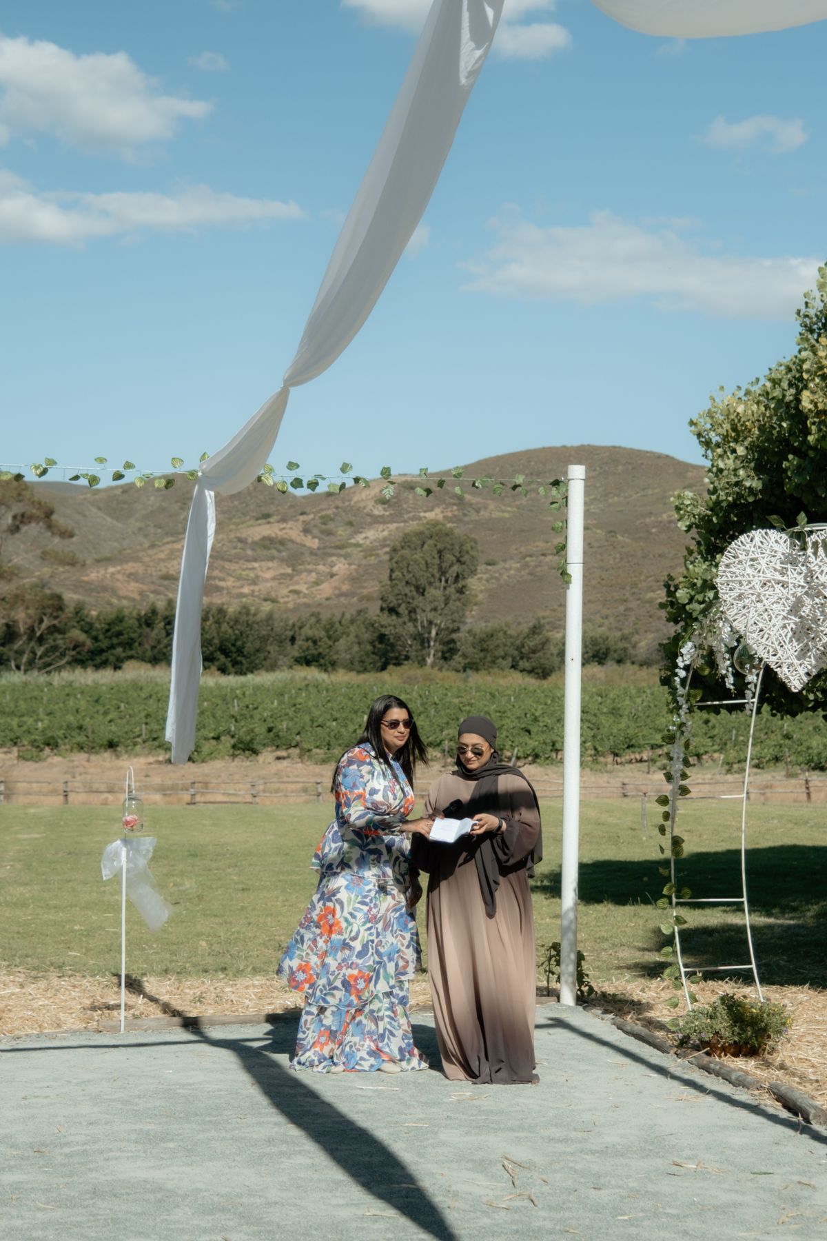 Two women in modest attire review documents at outdoor wedding ceremony with mountain views.