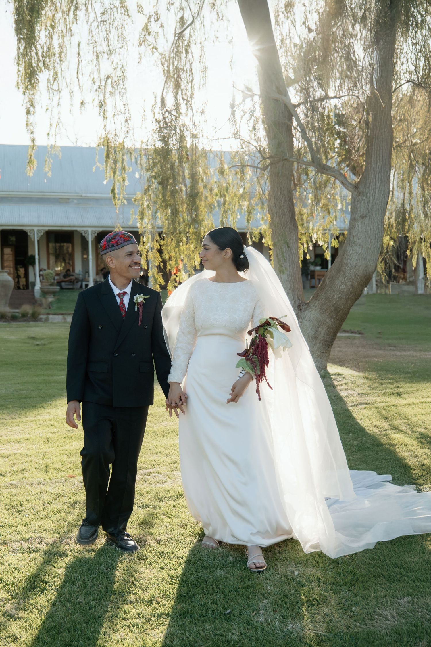 Muslim bride and groom walk hand-in-hand on green lawn beneath weeping willow tree during outdoor wedding ceremony.