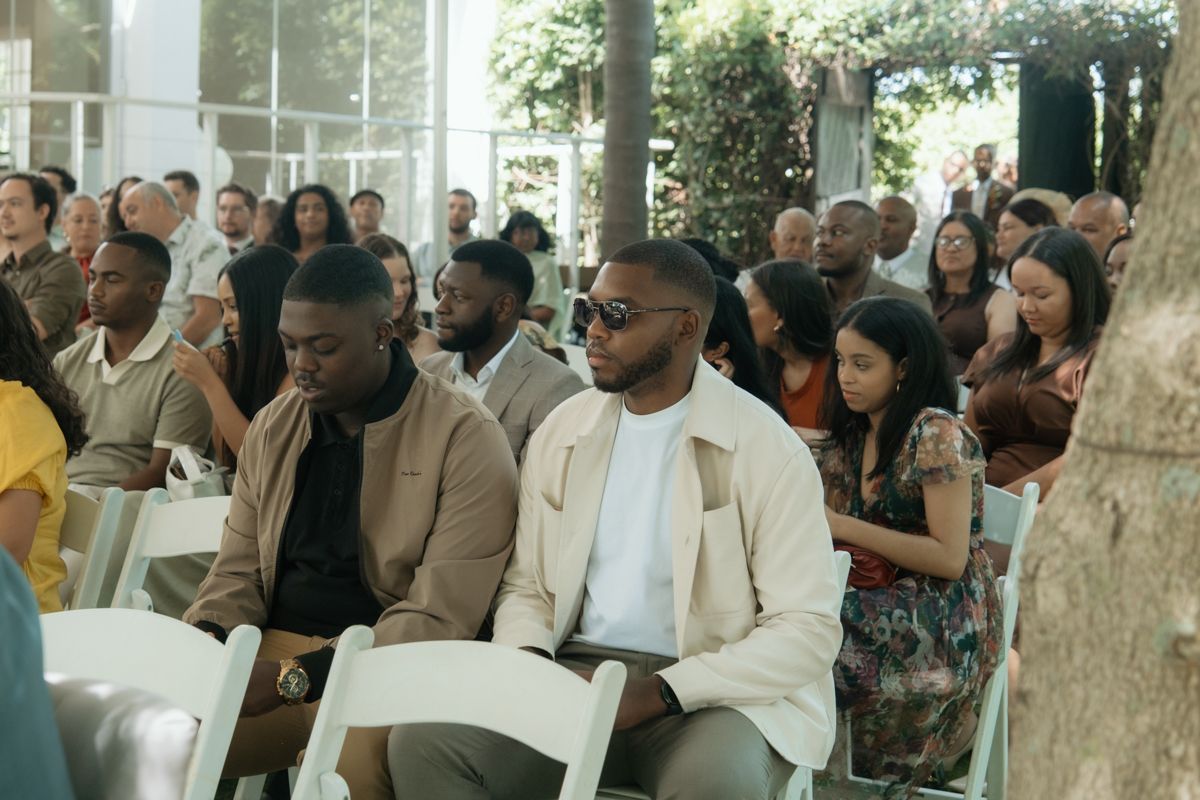 Diverse wedding guests seated in white chairs at an outdoor ceremony under trees.