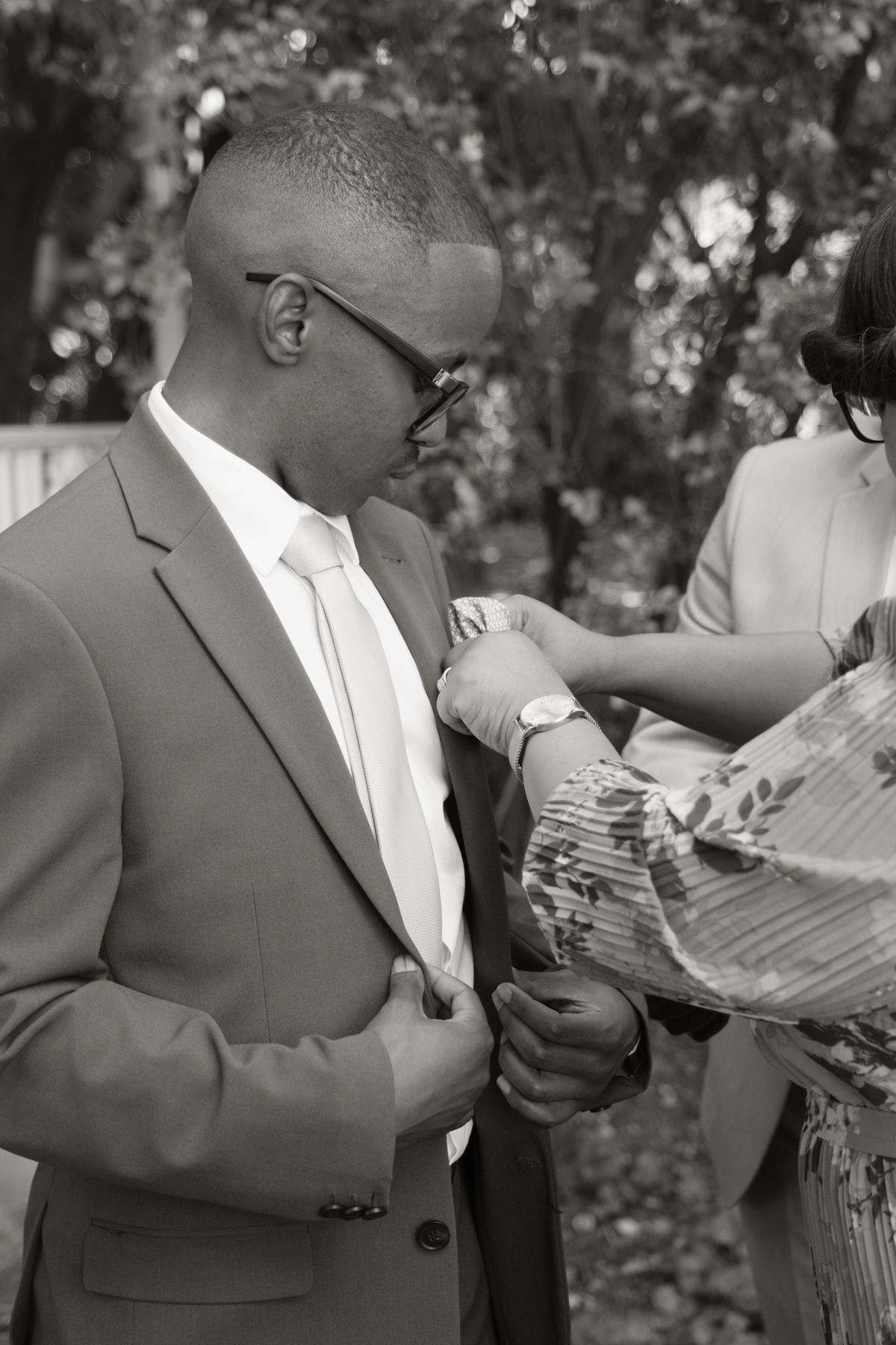 Black and white close-up of groom in tan suit having boutonniere pinned on lapel outdoors.