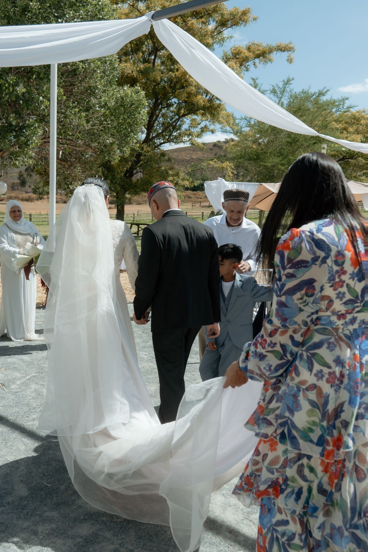 Bride in white gown and veil walking to outdoor wedding altar with groom and guests.