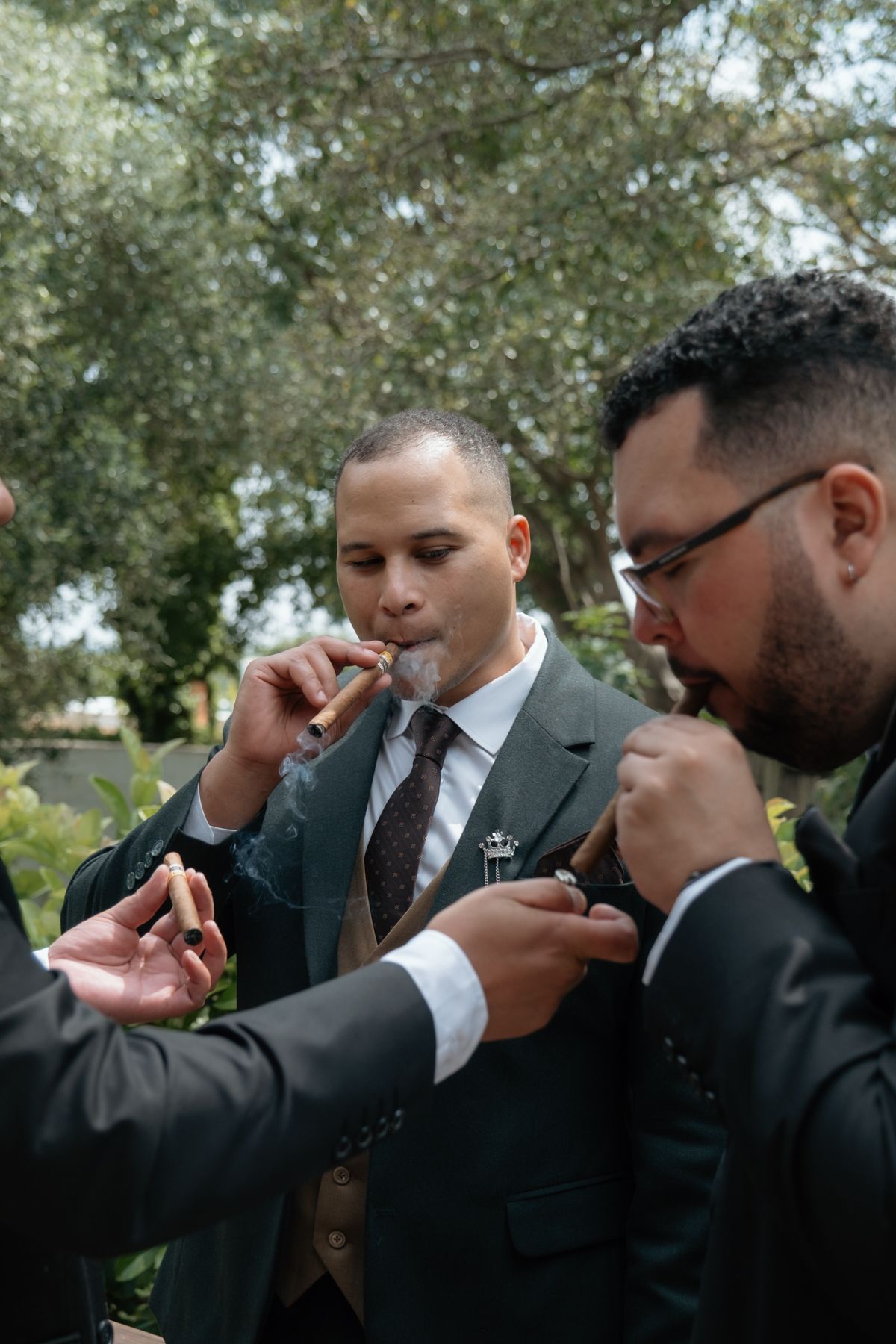 Groomsmen in suits lighting cigars outdoors before a wedding ceremony.