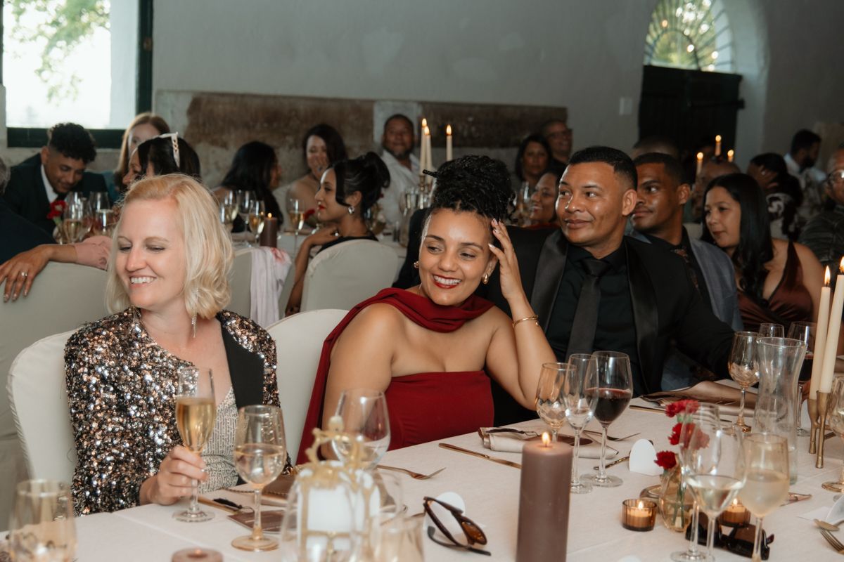 Wedding guests smile and laugh at reception table with wine glasses and candles in elegant venue.