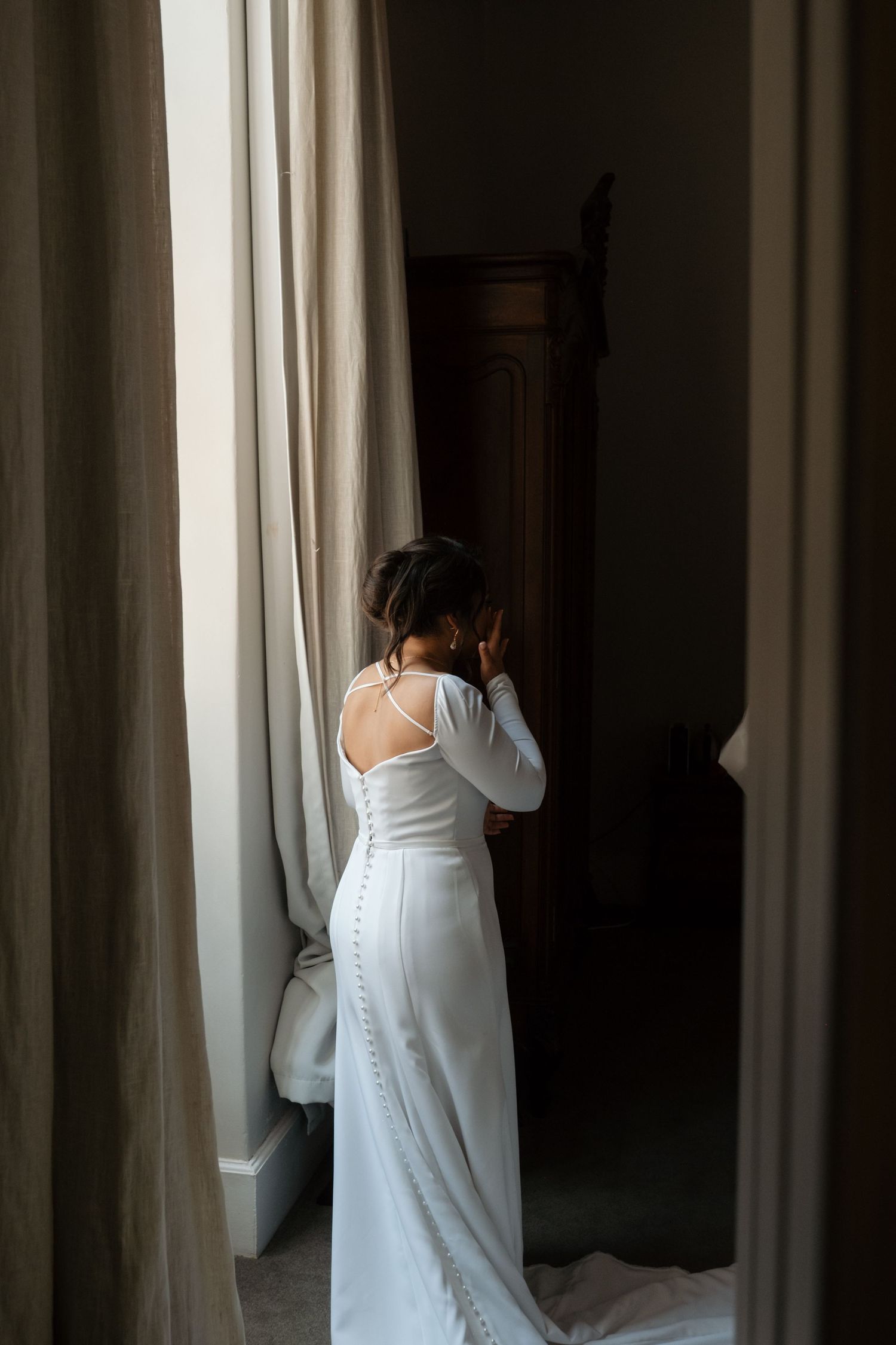 Bride in elegant white long-sleeve button-back wedding gown standing by curtained window.