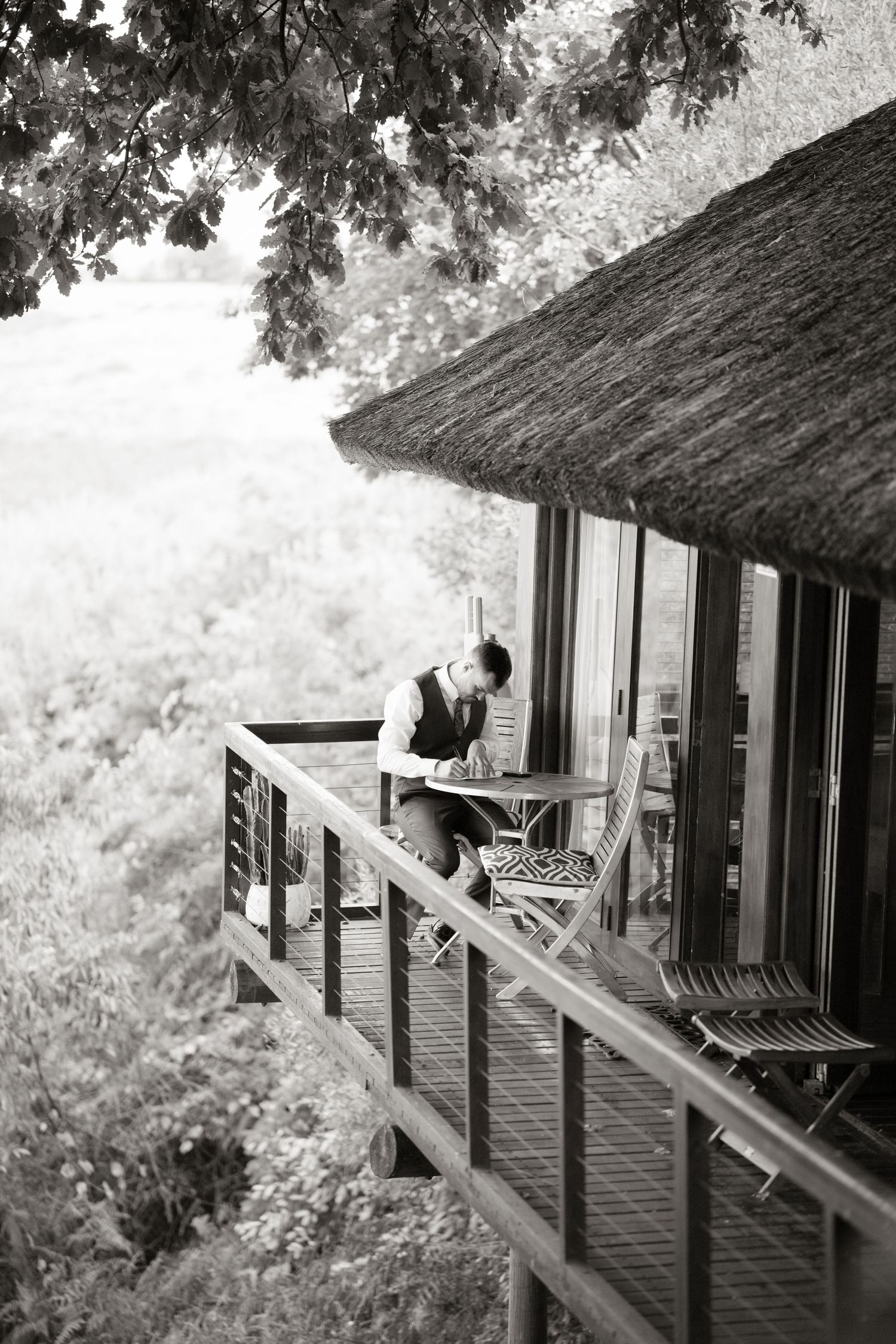 Man in vest writing at outdoor deck table overlooking lush forest landscape in black and white.