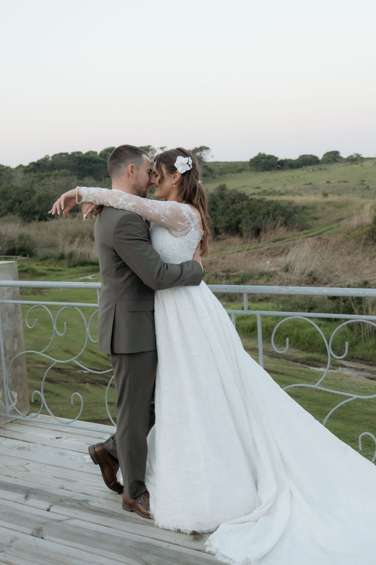 Bride in lace wedding gown and groom in gray suit embrace on scenic outdoor deck at dusk.