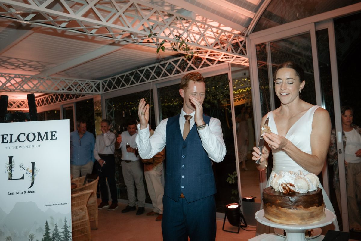 A groom in a navy vest stands beside a smiling bride holding a wedding cake at their reception.
