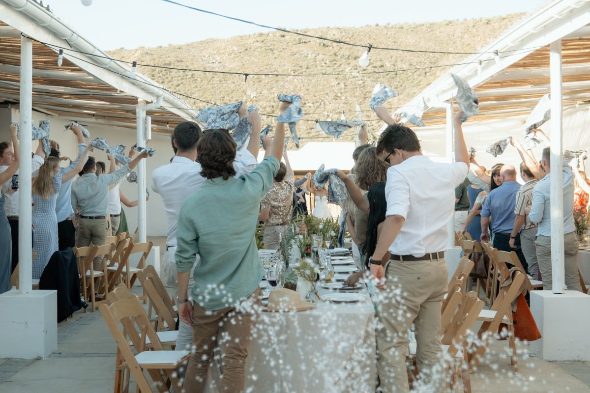 Guests celebrate at an outdoor wedding reception, raising their hands joyfully under a white pergola with floral decorations.