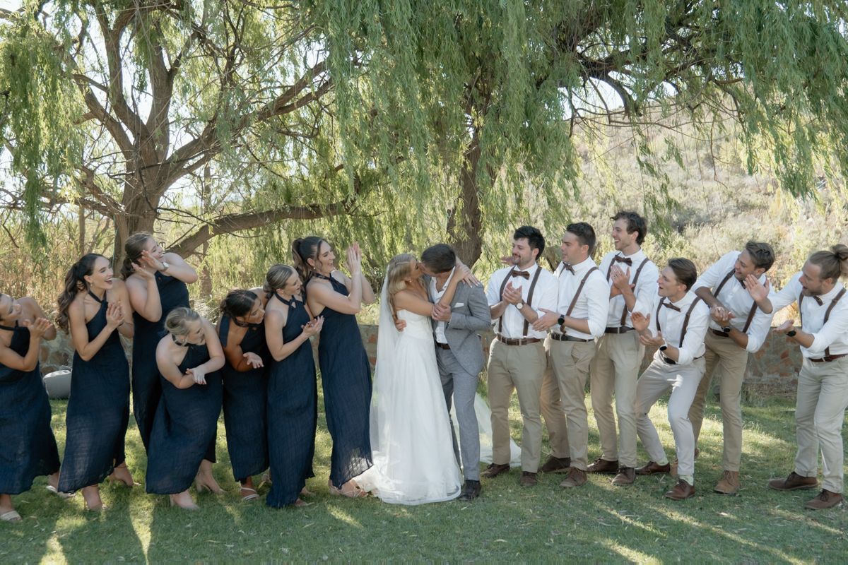 Wedding party laughing and celebrating under a weeping willow tree with bridesmaids in navy dresses and groomsmen in suspenders.