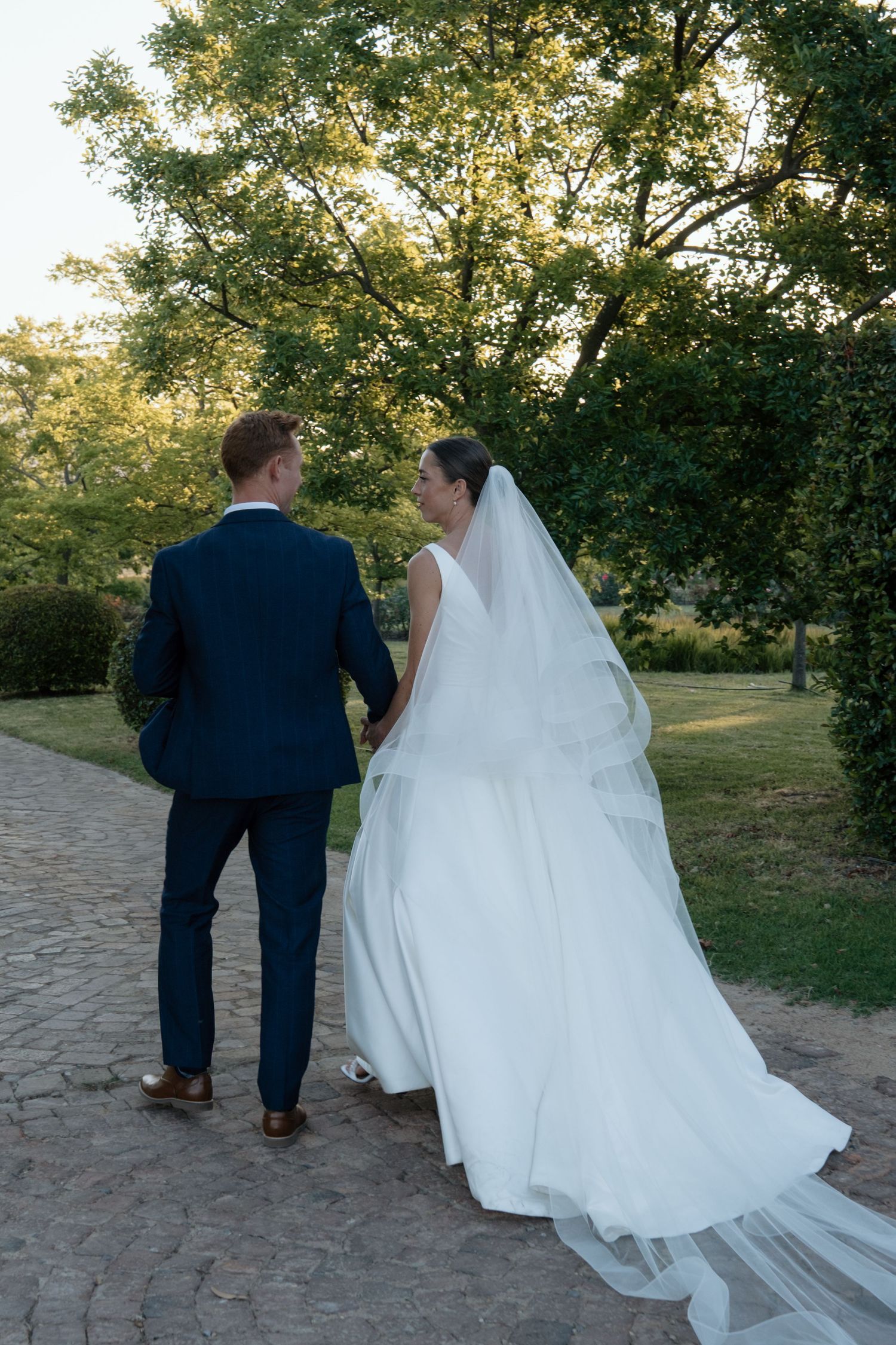 Bride and groom walk hand in hand through a lush green garden on their wedding day.