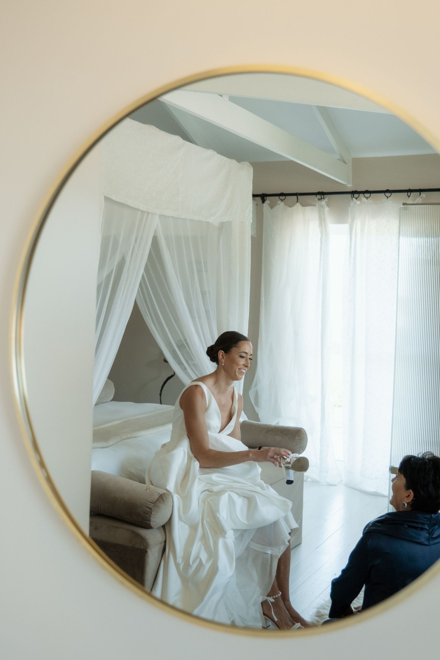 Bride in white gown smiles while seated, reflected in a round gold-framed mirror in an elegant bridal suite.