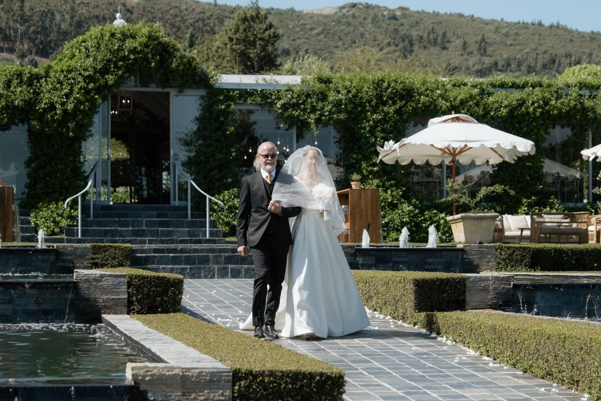 Bride in a white gown and veil walks with an older gentleman along a garden pathway at a sunny venue.