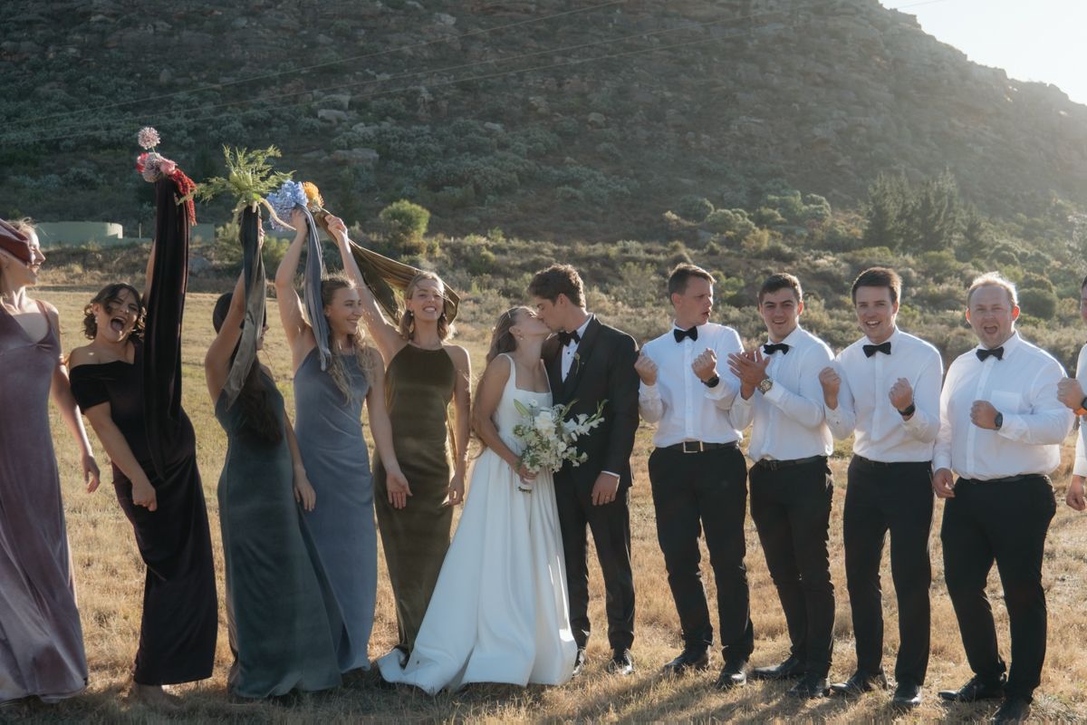 Wedding party celebrating outdoors with mountains backdrop, bride in white gown holding bouquet aloft with bridesmaids and groomsmen.