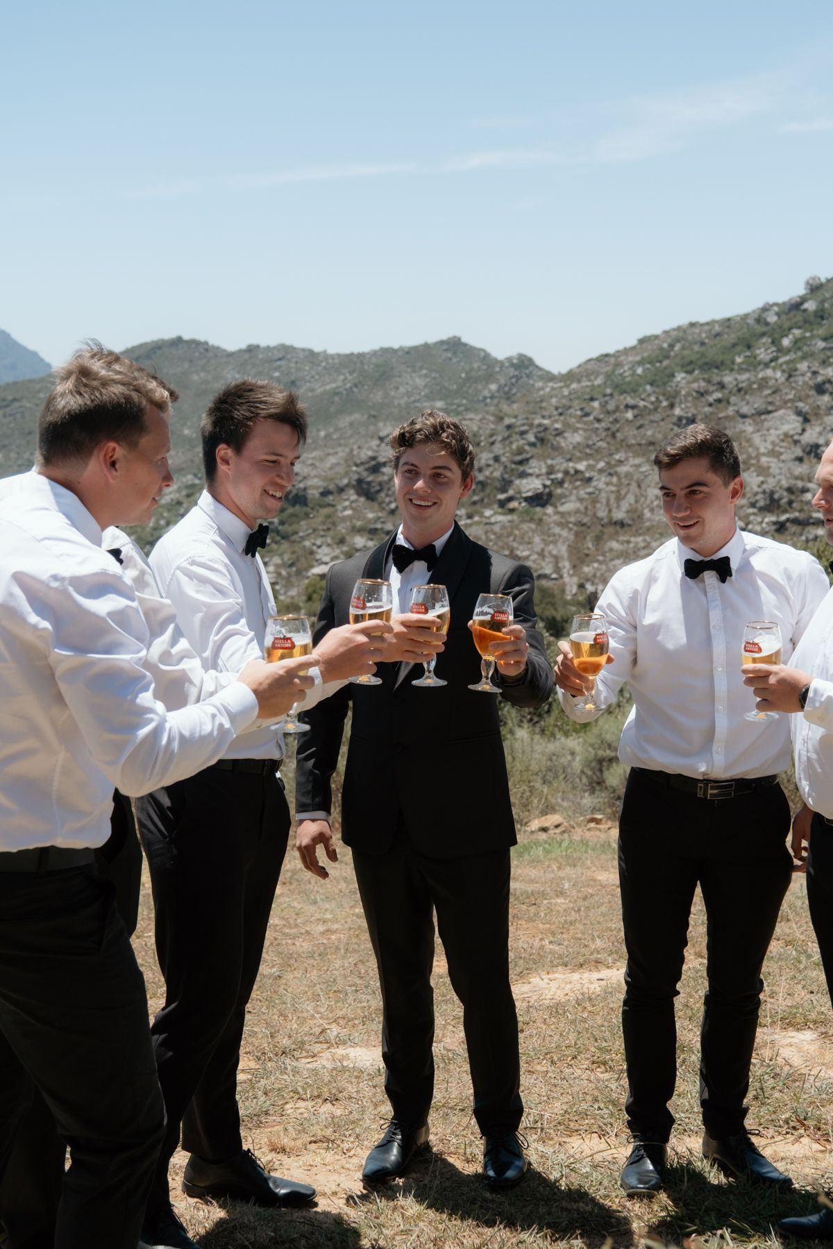 Groomsmen in black tuxedos and bow ties toasting with champagne glasses outdoors with mountain backdrop.