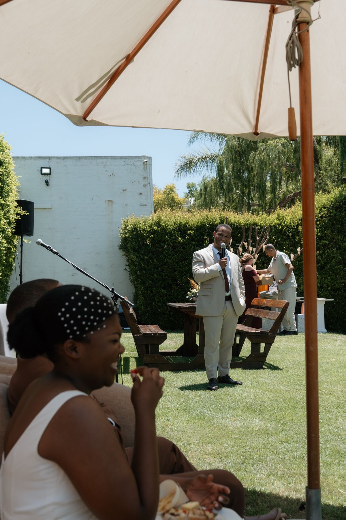 Man in suit speaks at outdoor garden event while guests sit under large patio umbrella on sunny day.