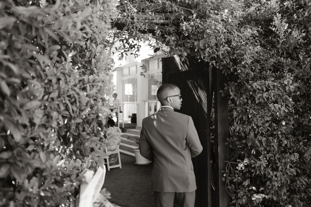Black and white photo of a man in a suit walking through a garden archway at an outdoor wedding venue.