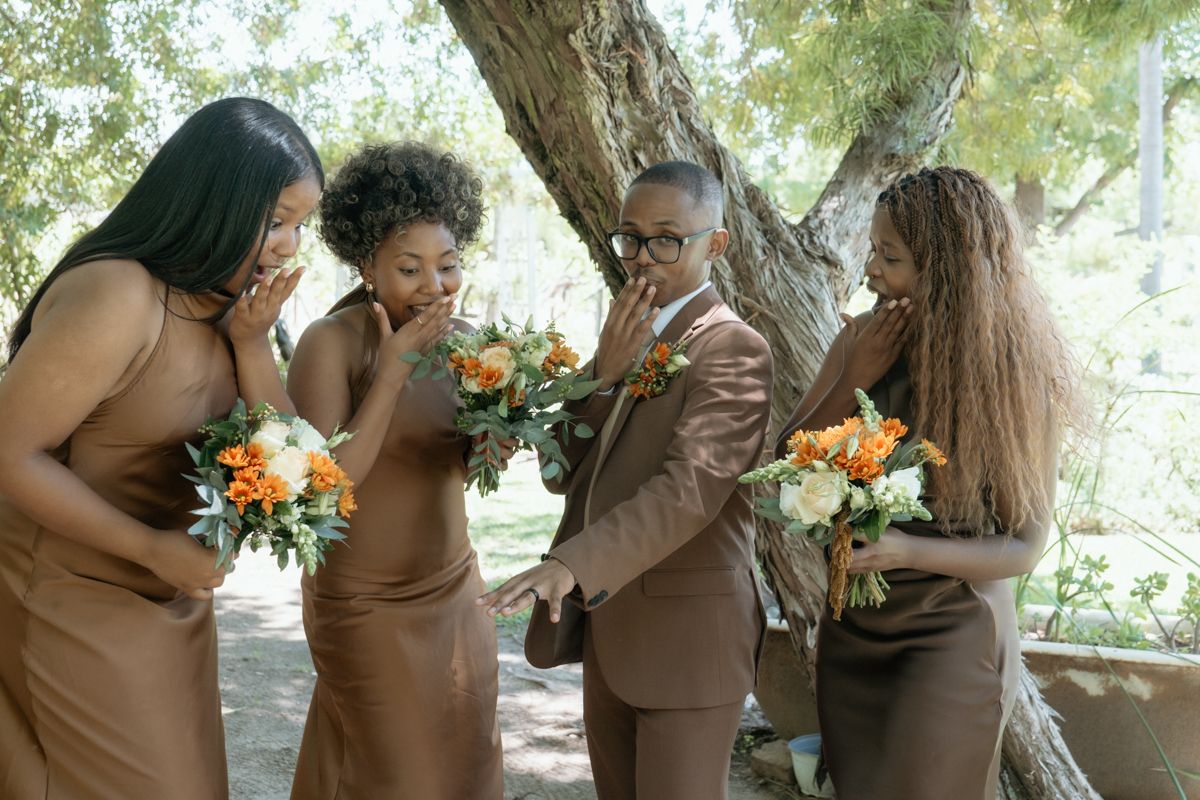 Groom in brown suit poses with three bridesmaids in matching brown dresses holding orange and white bouquets outside.