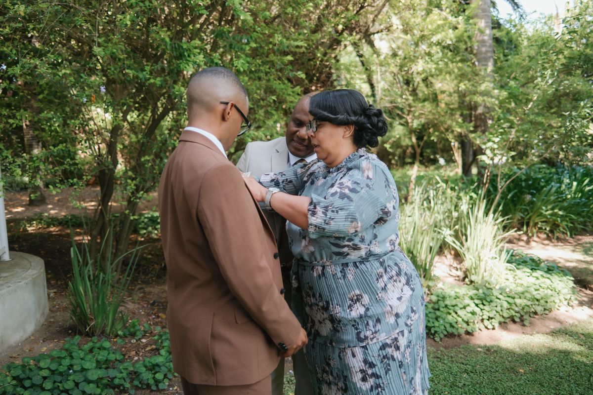 A groom in a brown suit receives a boutonniere from a woman in a floral dress in a garden setting.