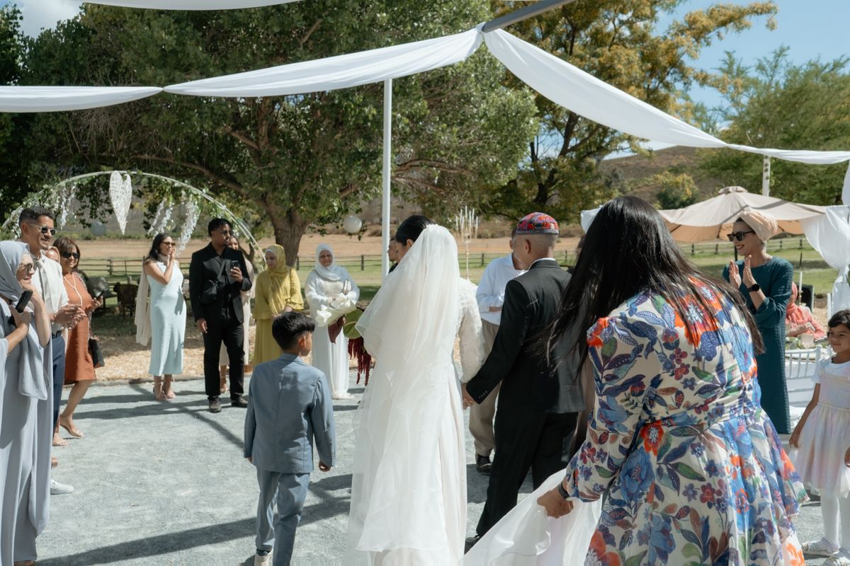 Wedding procession with bride in white veil walking toward outdoor ceremony under draped canopy.