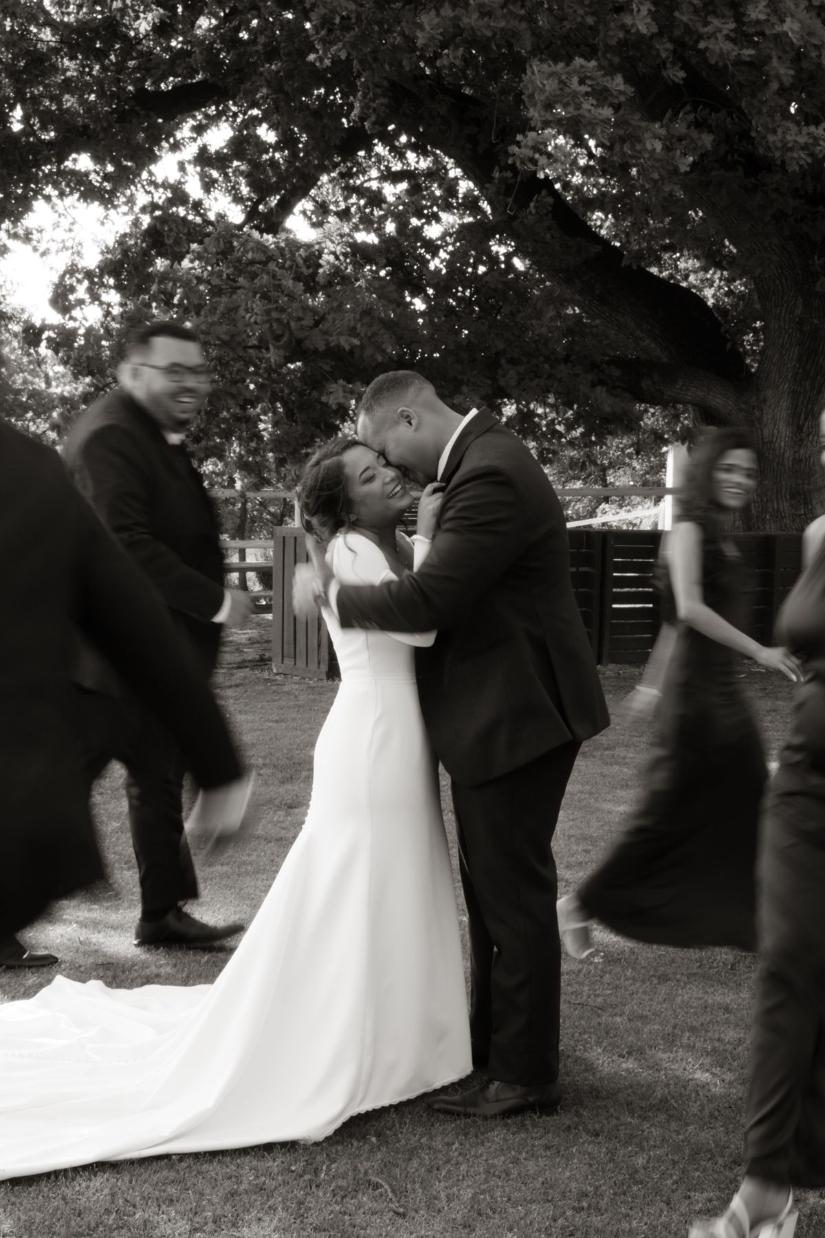Black and white photo of newlyweds embracing while wedding guests move around them outdoors.